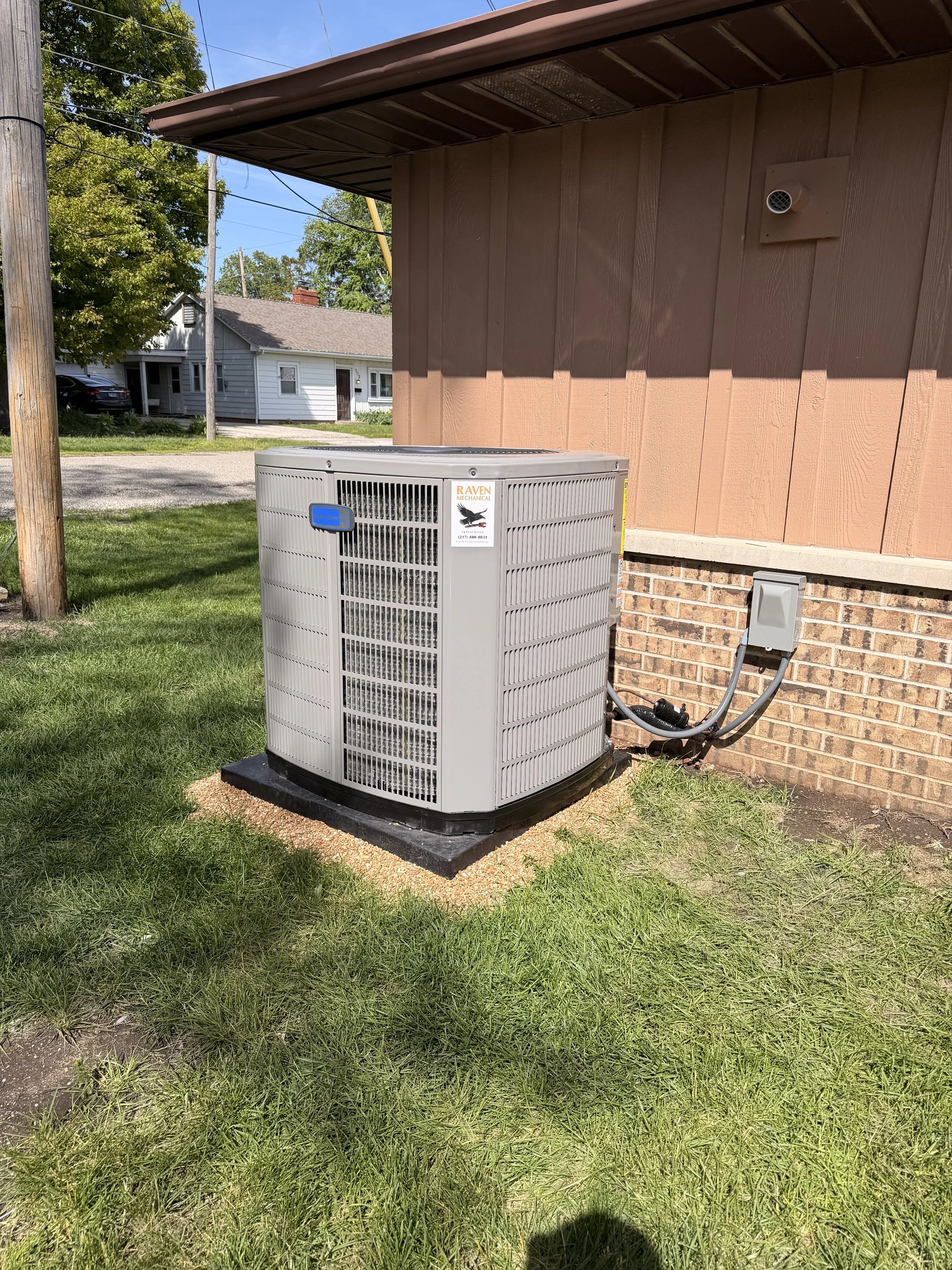 An outdoor air conditioning unit installed on a concrete pad next to a house with brown siding and brick foundation, on a grassy lawn under a small roof extension.