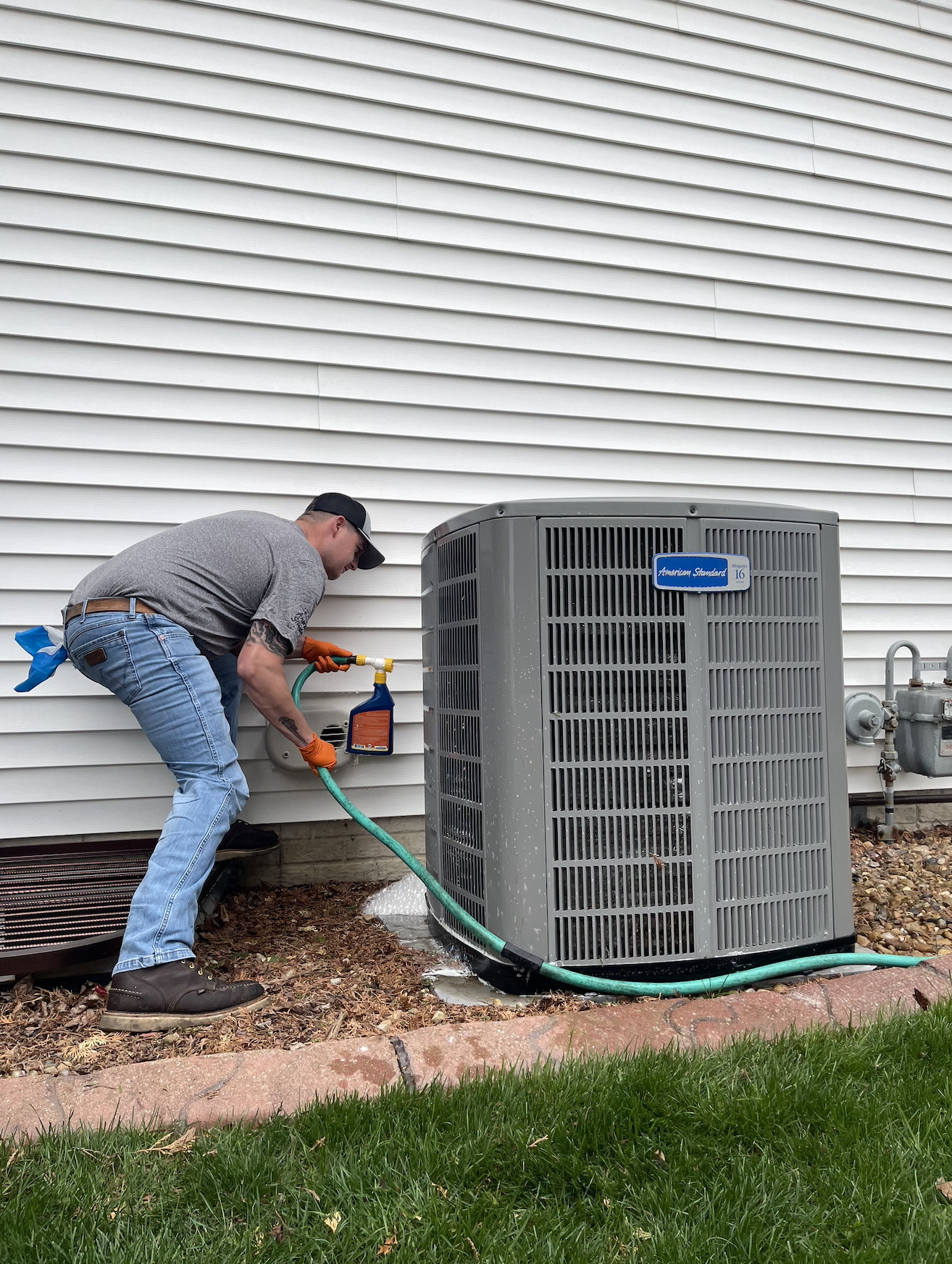 A man wearing a black cap, gray t-shirt, and blue jeans, with orange gloves, is cleaning an outdoor air conditioning unit with a spray bottle.