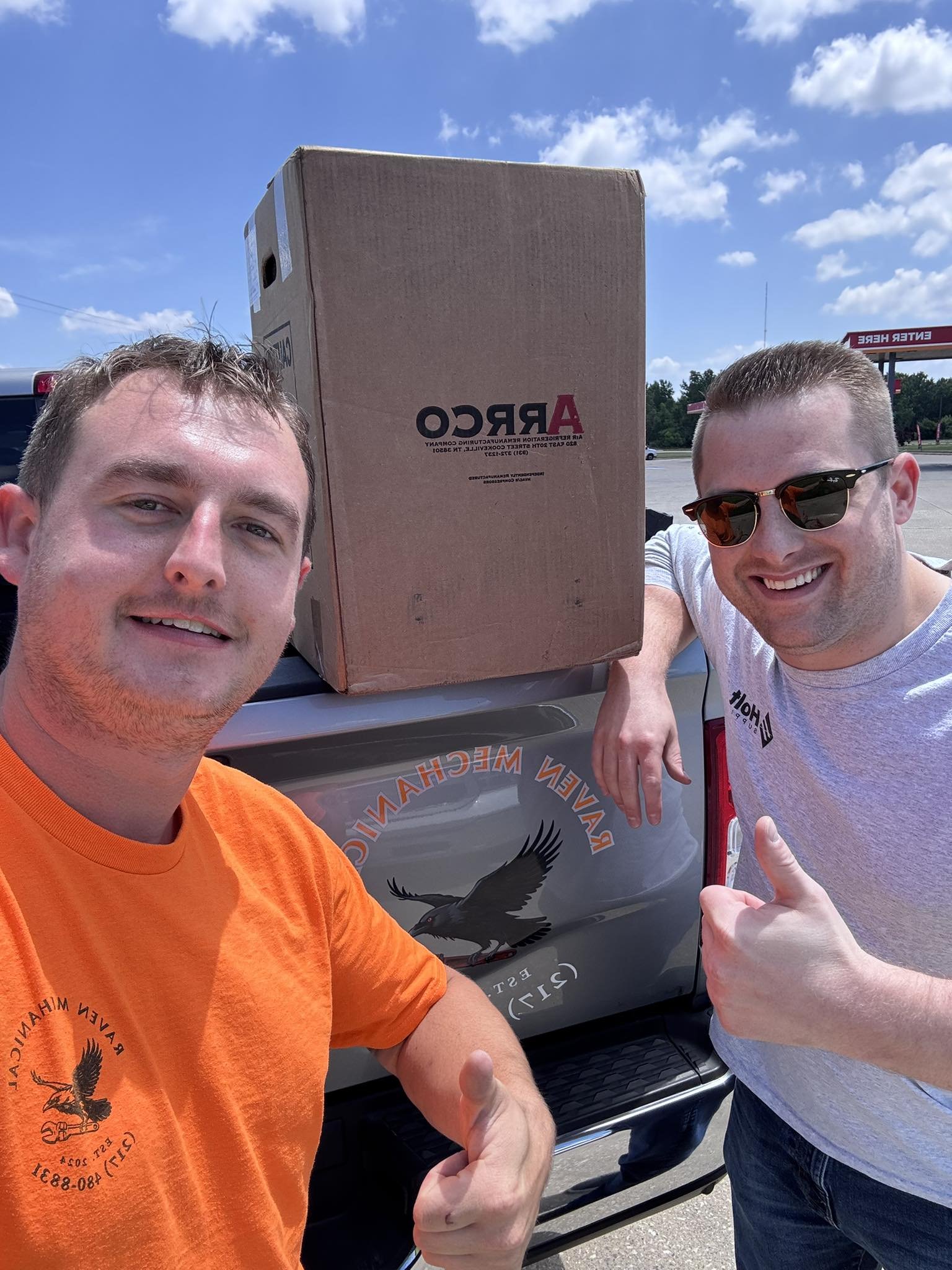 Two smiling men taking a selfie outdoors near a truck with an eagle logo and a large cardboard box, blue sky with some clouds in the background.