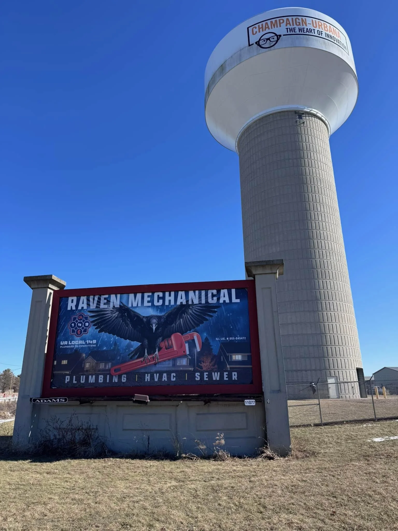 A water tower with a round top and a tall cylindrical base, with a sign in front advertising Raven Mechanical services including plumbing, HVAC, and sewer.