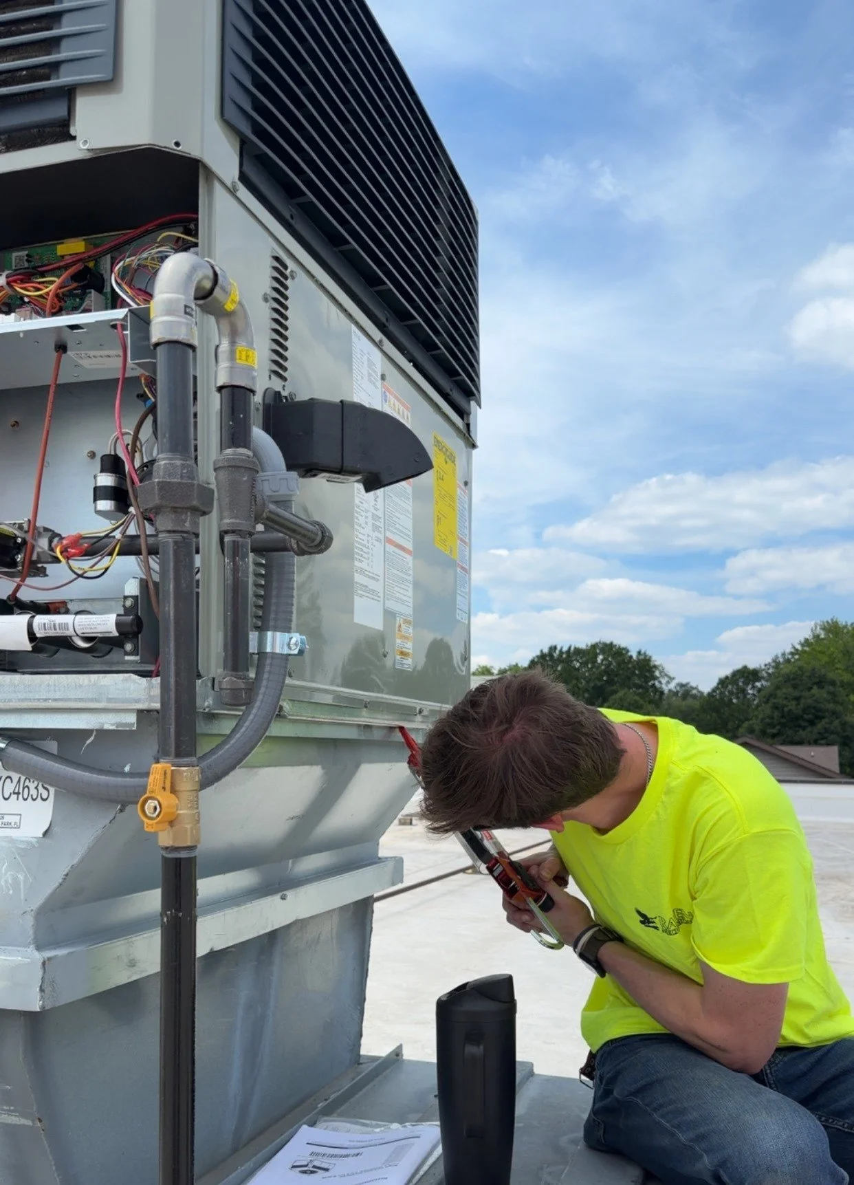 Technician working on an HVAC unit outdoors, inspecting and repairing the equipment with tools, with a blue sky and trees in the background.