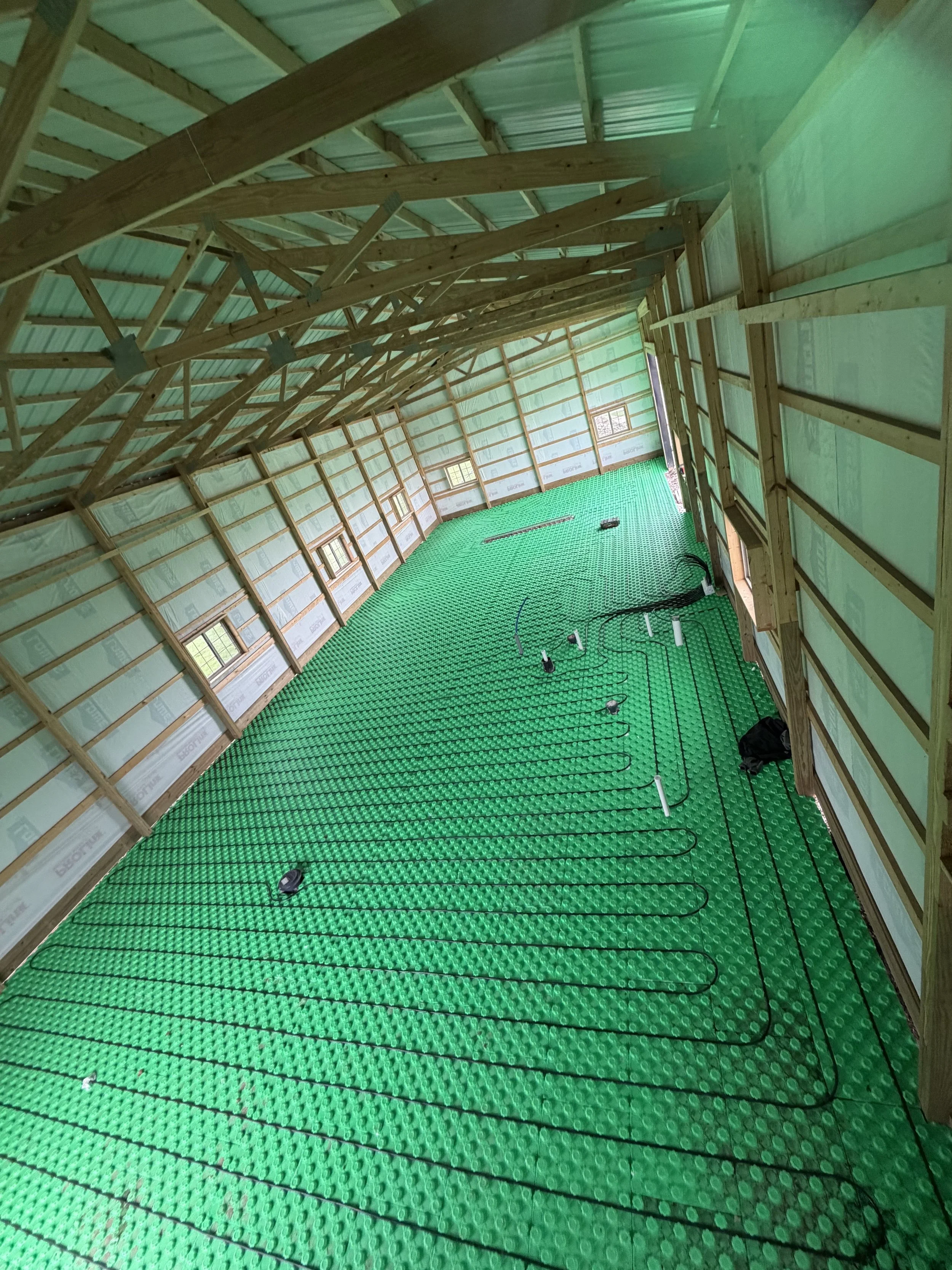 Interior of a building under construction, showing wooden framing and a green radiant floor heating system installed on the floor.