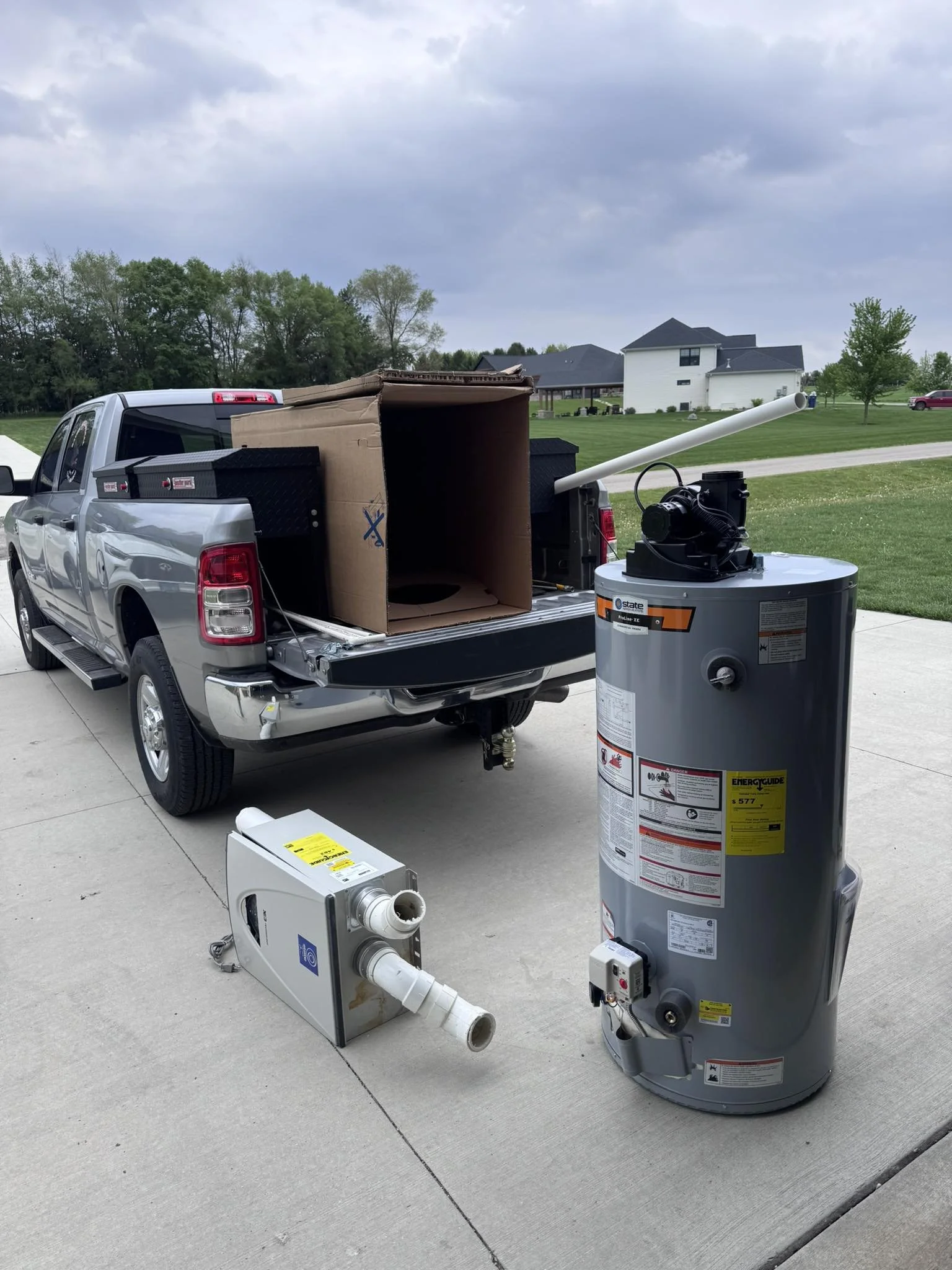 A pickup truck parked on a driveway with a water heater and ducting materials set up nearby, against a backdrop of green lawn, trees, and a large house, with a cloudy sky overhead.