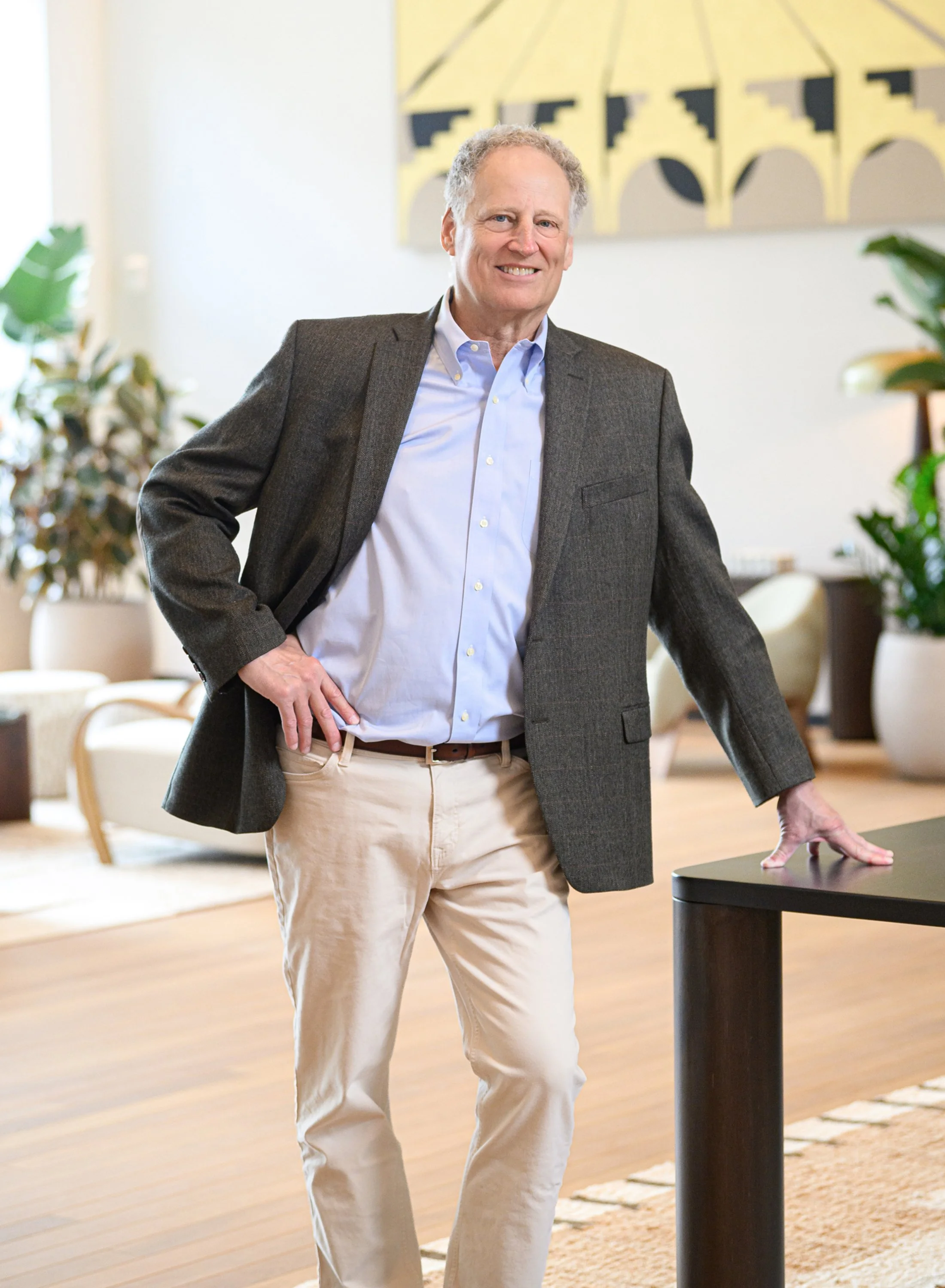 A smiling middle-aged man with curly gray hair wearing a gray blazer, light blue dress shirt, and beige pants standing in a modern, well-lit room with plants and abstract artwork on the wall.