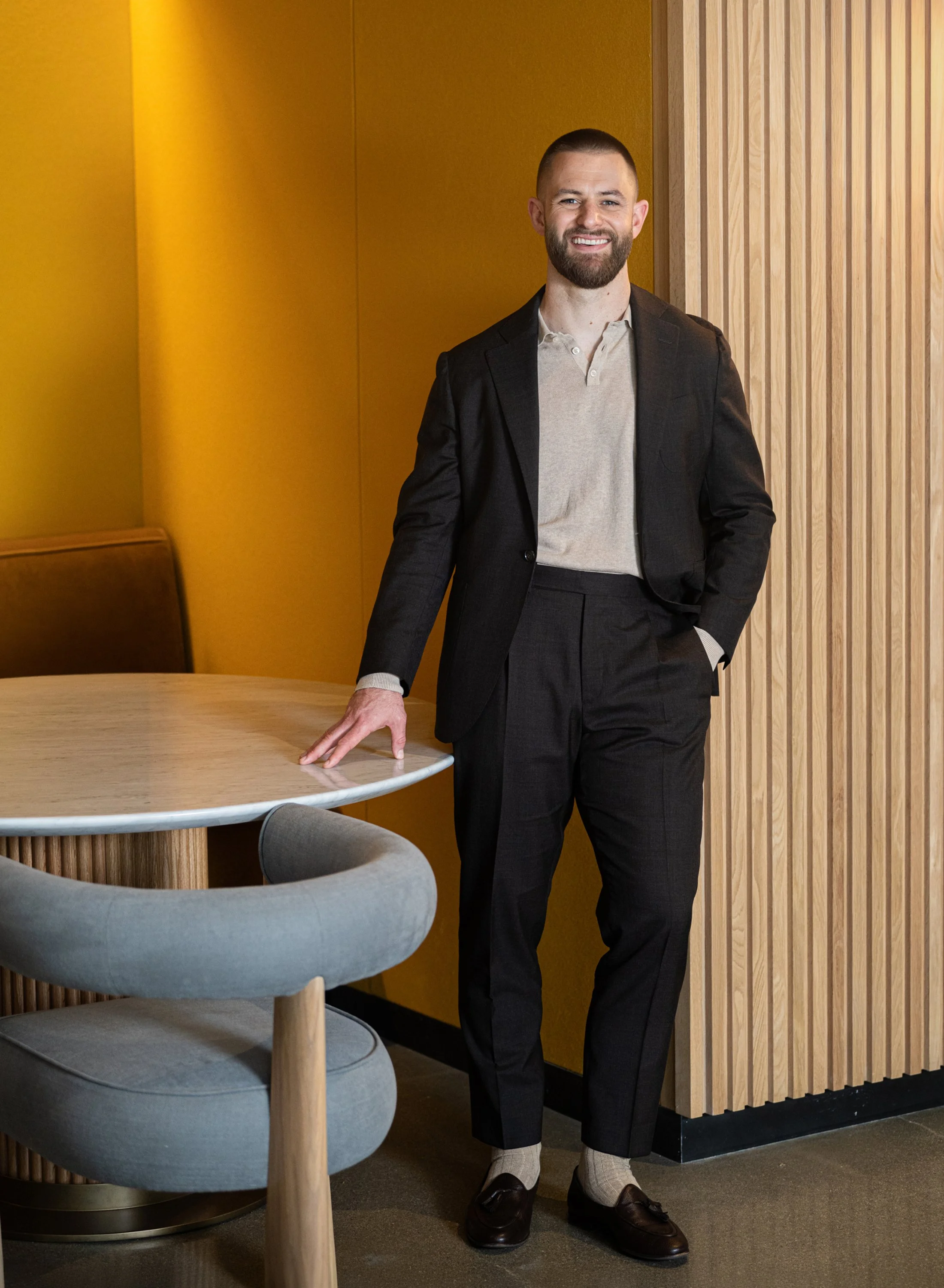 A man with a beard in a suit standing next to a table in a room with wooden wall paneling and yellow paint.