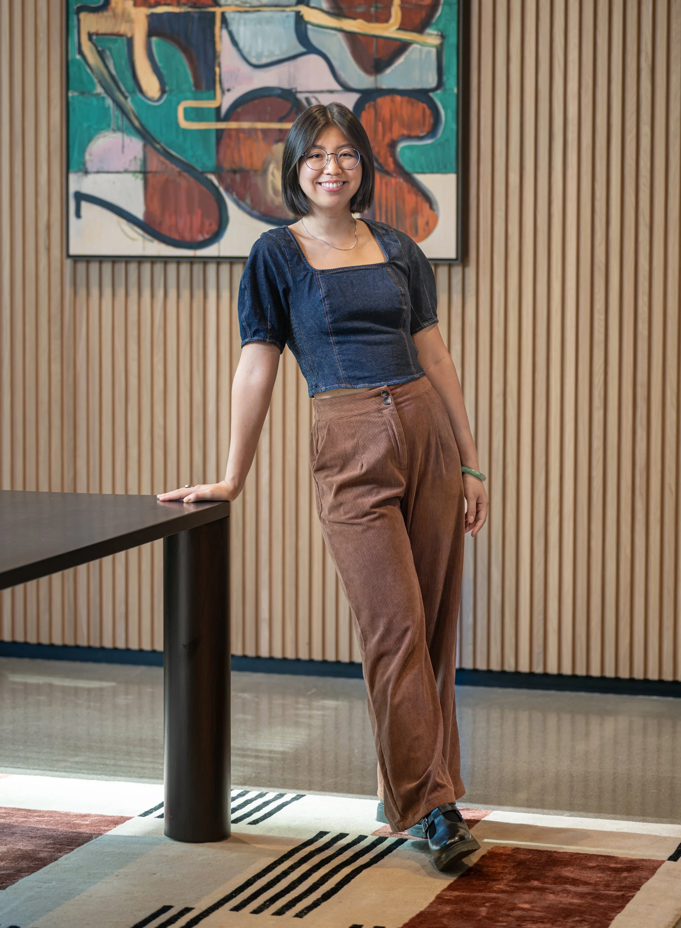 A young woman with shoulder-length dark hair, glasses, and a wide smile, standing indoors in front of a colorful abstract painting and wooden-paneled wall. She is wearing a dark denim crop top, brown pants, and black shoes, with her right hand resting on a black table.