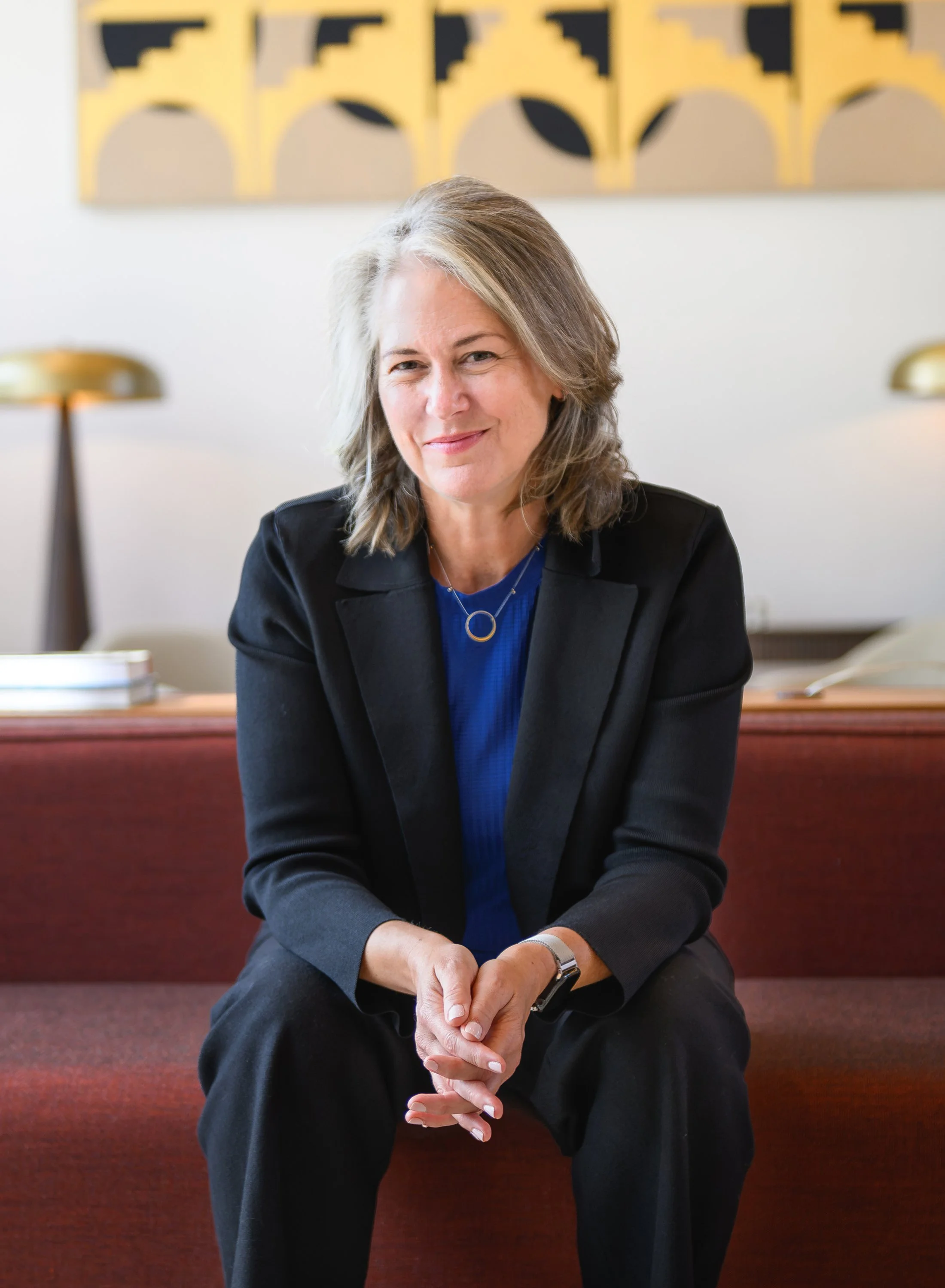 A woman with shoulder-length gray hair wearing a black blazer and blue top, sitting on a red upholstered bench, smiling at the camera in a room with modern artwork on the wall and two gold-colored lamps.
