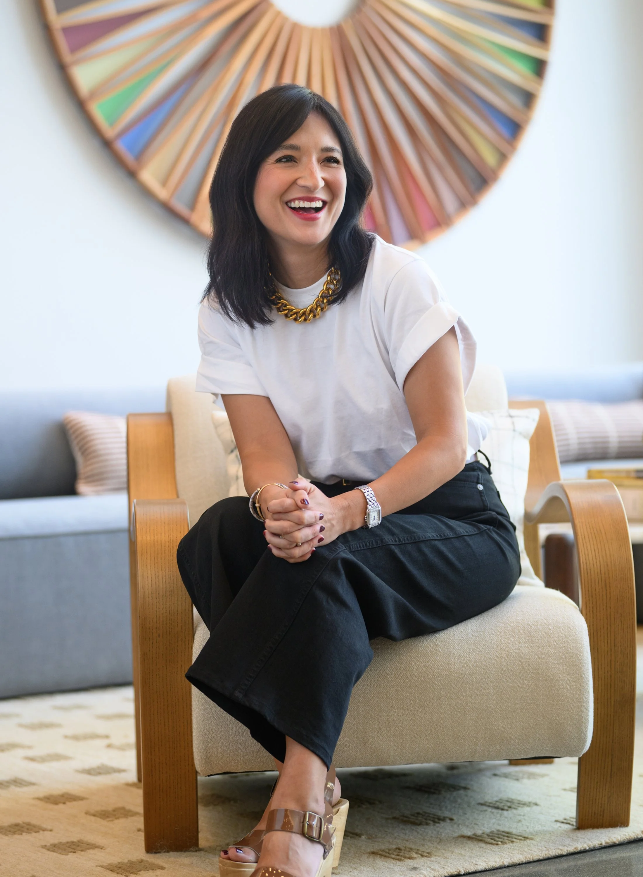 A woman with shoulder-length black hair sitting on a beige armchair in a modern room. She is wearing a white T-shirt, black pants, transparent wedge sandals, a gold chain necklace, a silver watch, and bracelets. She is smiling and looking to her right. Behind her is a decorative wall art piece with a circular, colorful, geometric design.
