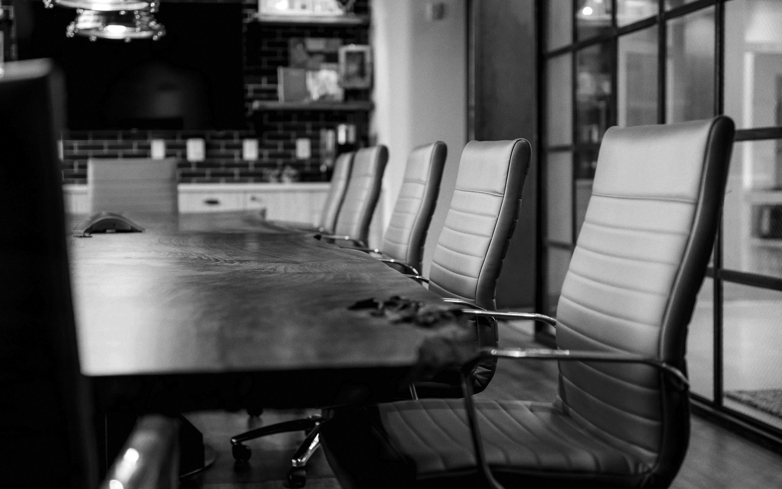 A black-and-white photo of a conference room with empty high-backed chairs arranged around a large wooden table. The room features glass panels on one side and a brick wall with decorative items in the background.