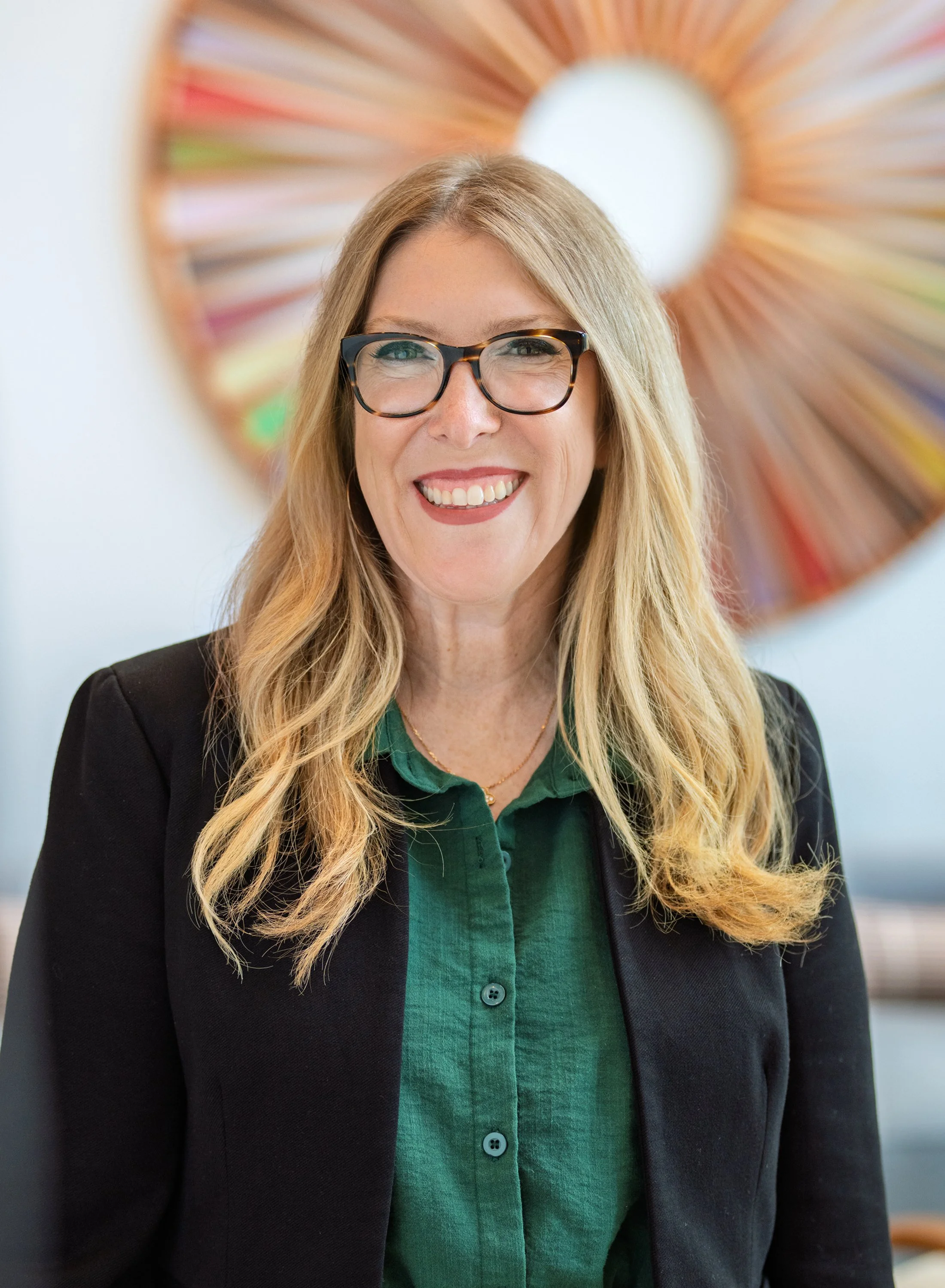 A smiling woman with long blonde hair, wearing glasses, a black blazer, and a green blouse, standing in front of a colorful circular art piece.