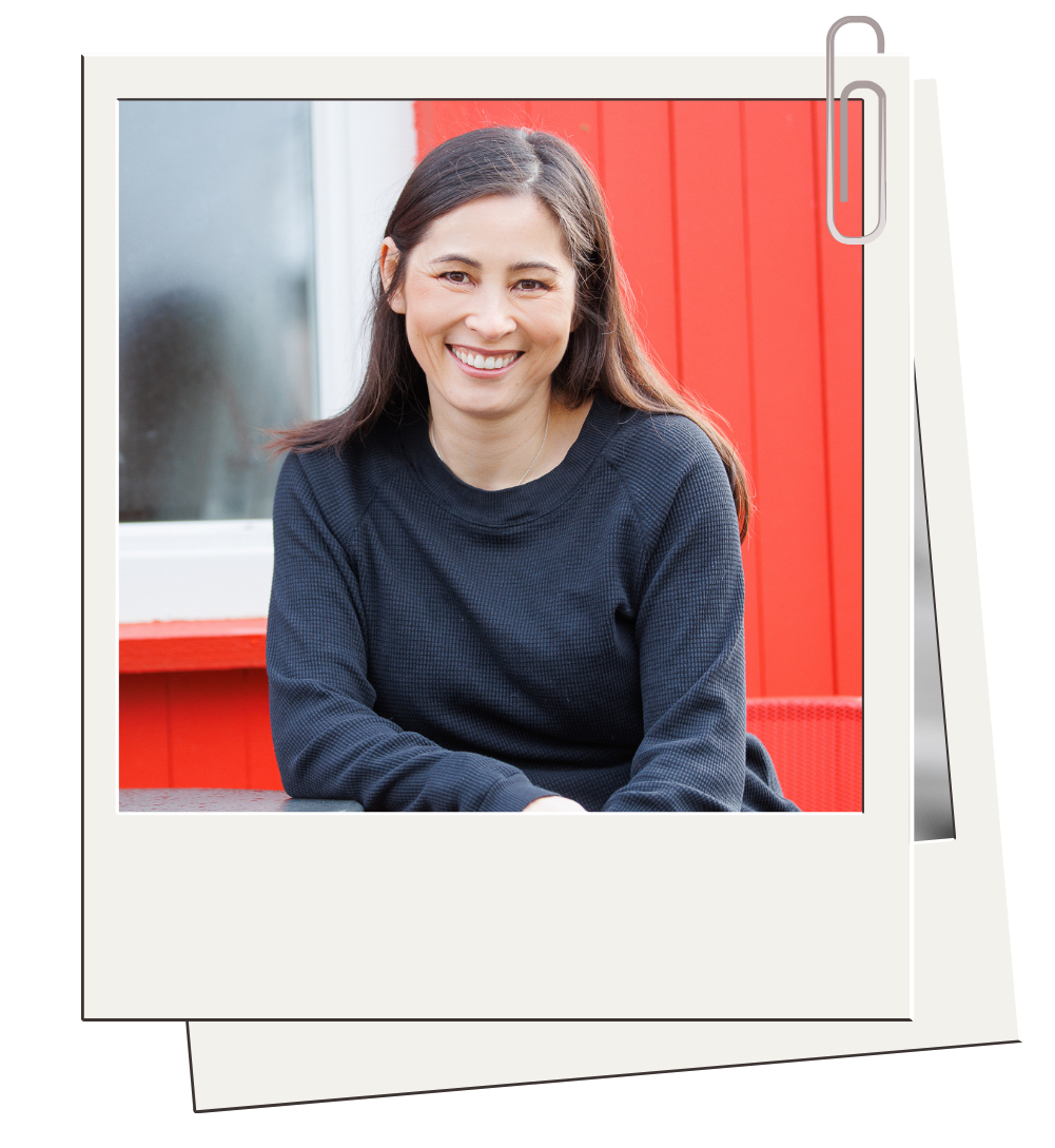 A woman with long brown hair smiling, wearing a dark blue sweater, sitting outdoors with a red wall and window in the background.