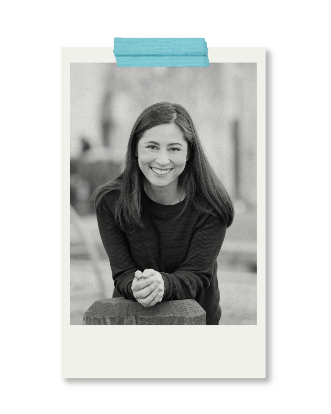 Black and white photo of a smiling woman with long hair, leaning on a wooden surface outdoors.