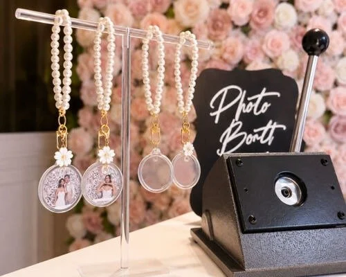 Jewelry hanging on a rack at a photo booth near a black sign with white cursive writing and a black camera box in front, with pink flowers in the background.