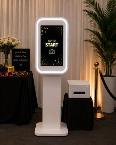 Touch screen kiosk with illuminated border displaying 'Tap to Start', situated indoors near a black table with a sign, glasses, and flowers, and next to a potted plant.