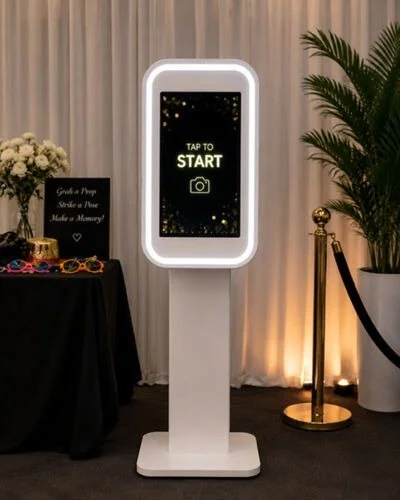 Photo of a photo booth stand with a touchscreen display that reads "Tap to Start" and a camera icon, set against a backdrop of white curtains and indoor plants.