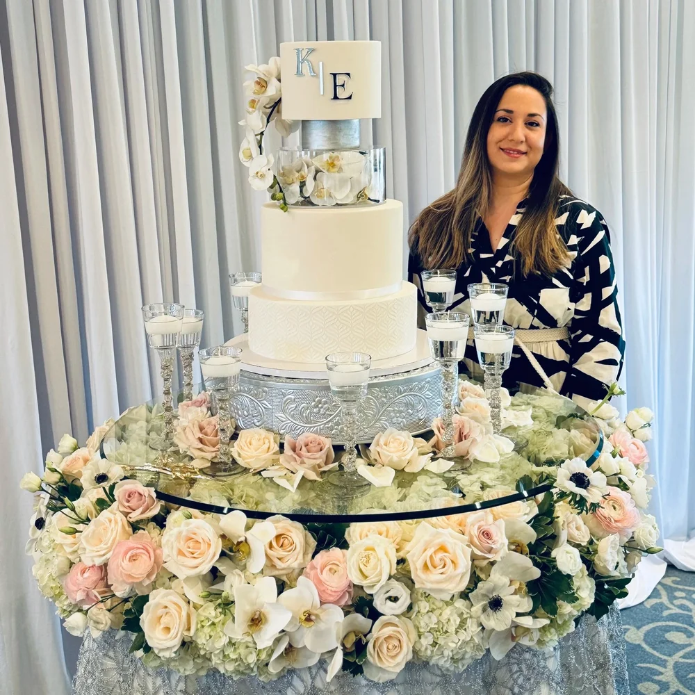 Chef Tina beside a tiered cake and an arrangement of pale flowers.