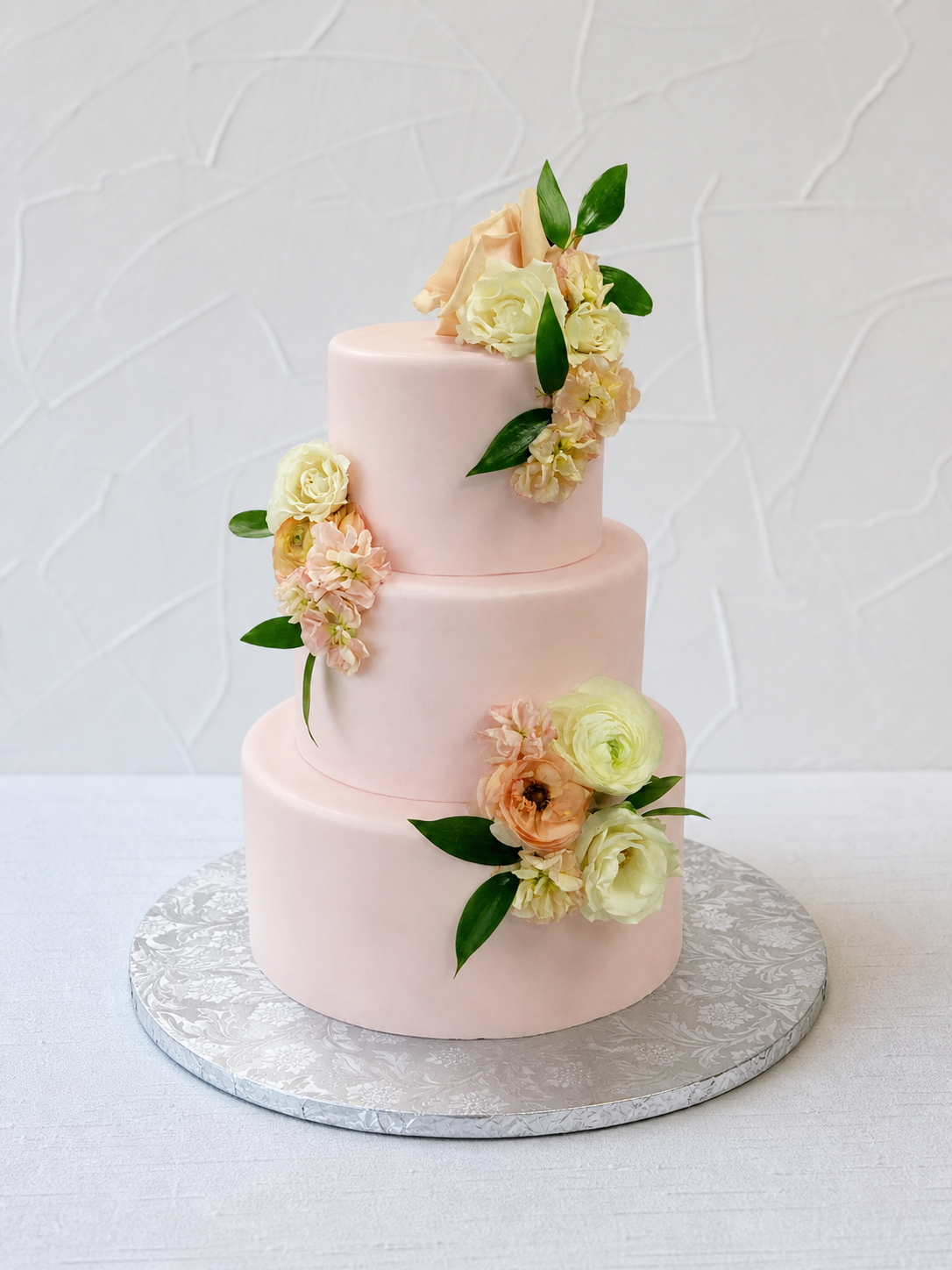 Pale pink tiered cake with peach and cream flowers and green leaves.