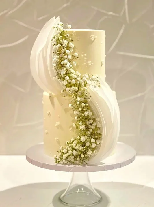 White tiered cake with flowing white decorations and small white flowers on a glass pedestal.