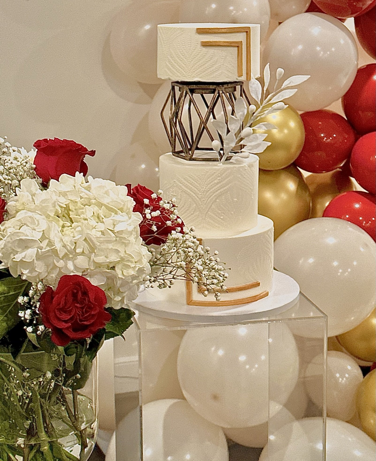 White and gold tiered cake with clear sections, displayed among red and gold decorations.