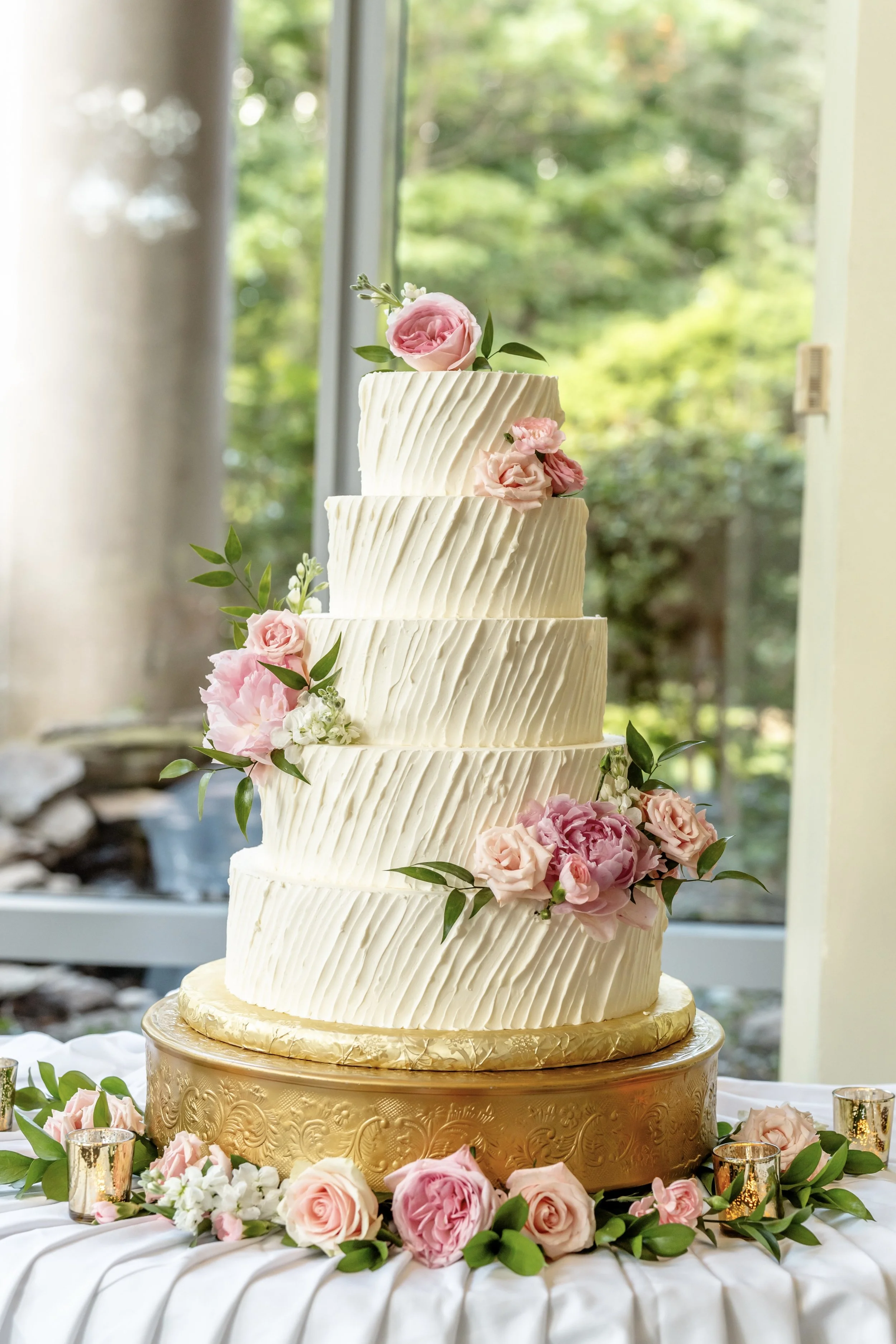 White tiered cake with textured icing and pink flowers on a gold stand.