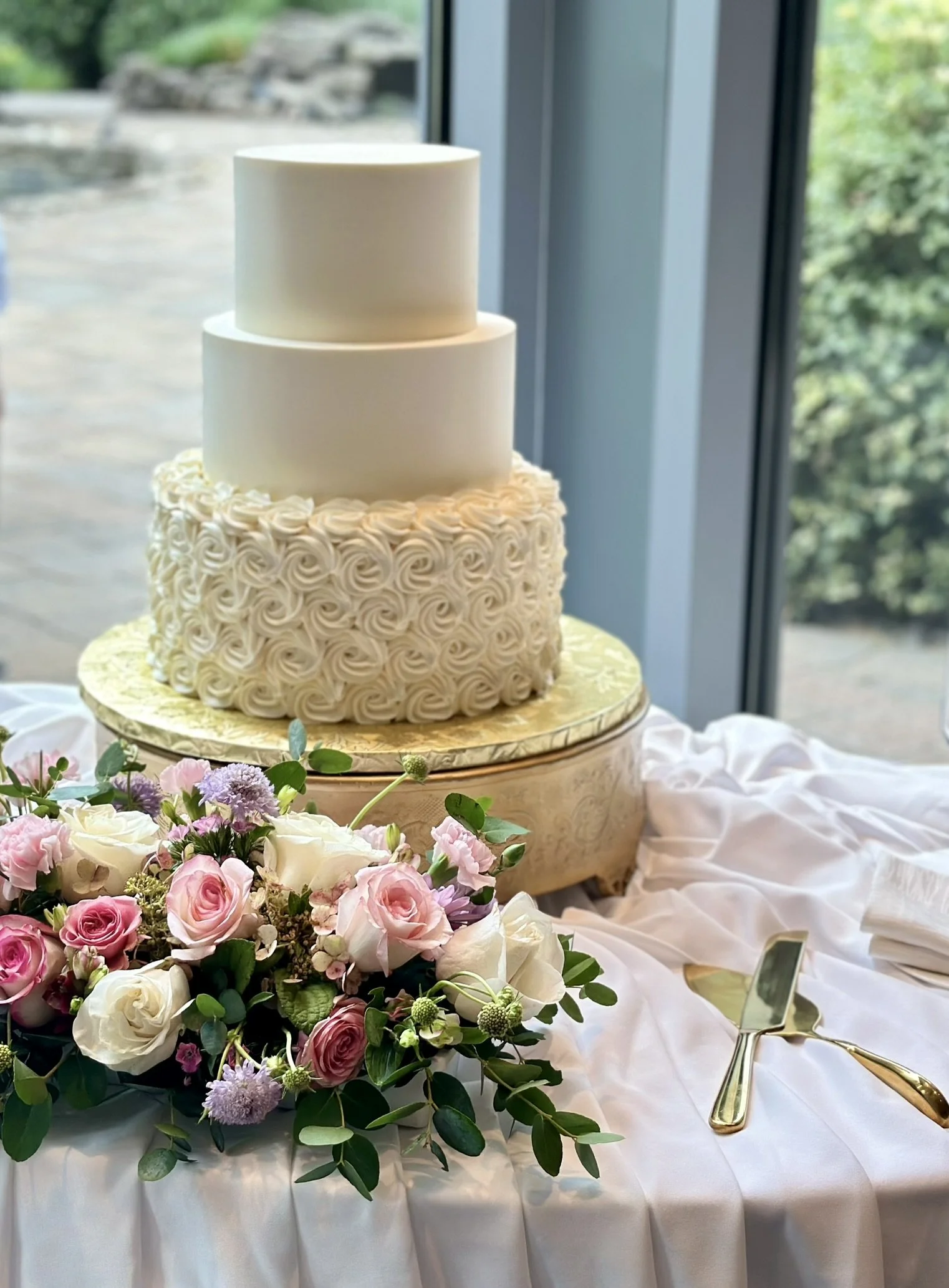 White tiered cake with a textured lower tier, displayed beside pink roses.