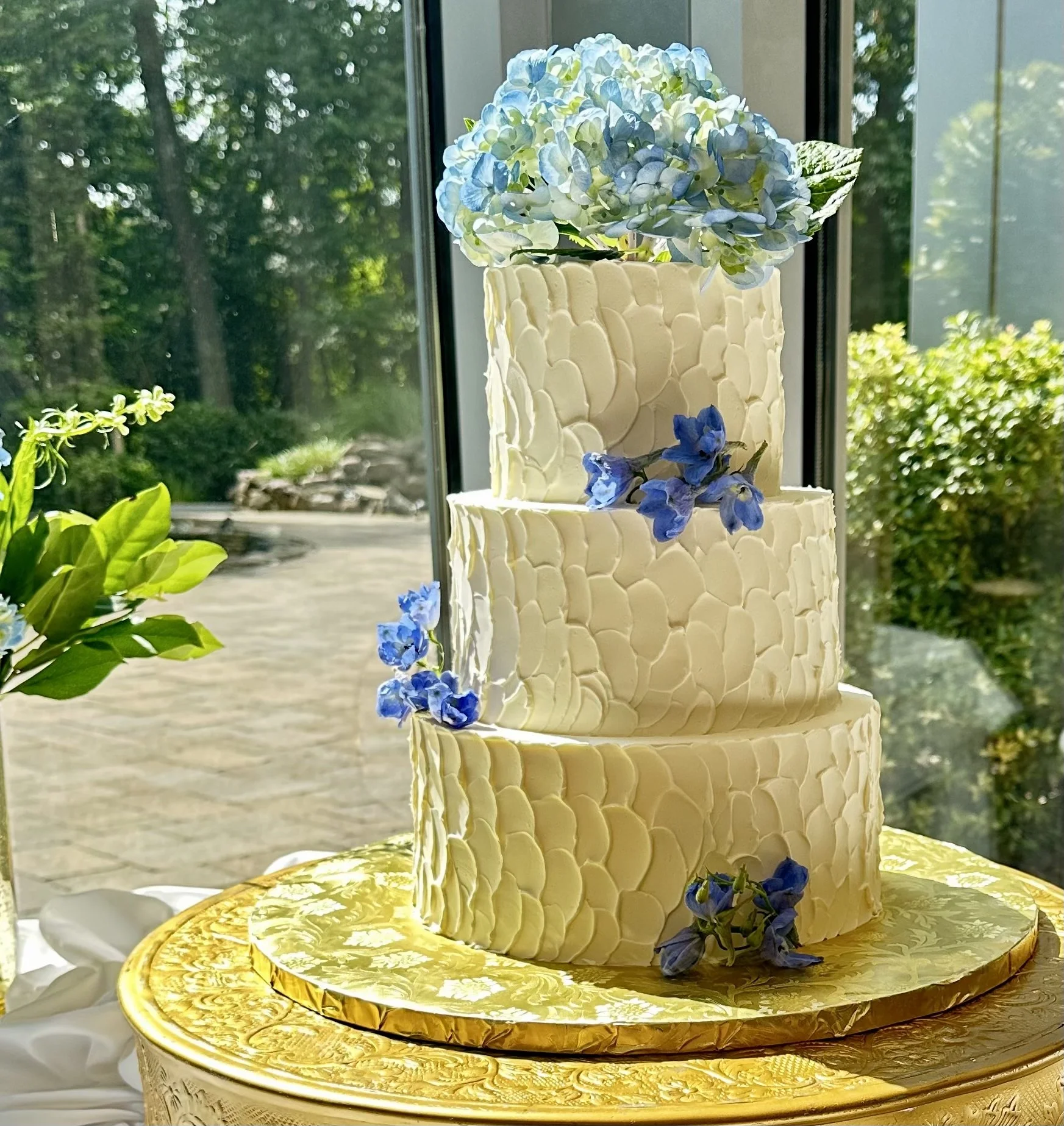 White and gold tiered cake with a clear central section and blue flowers.