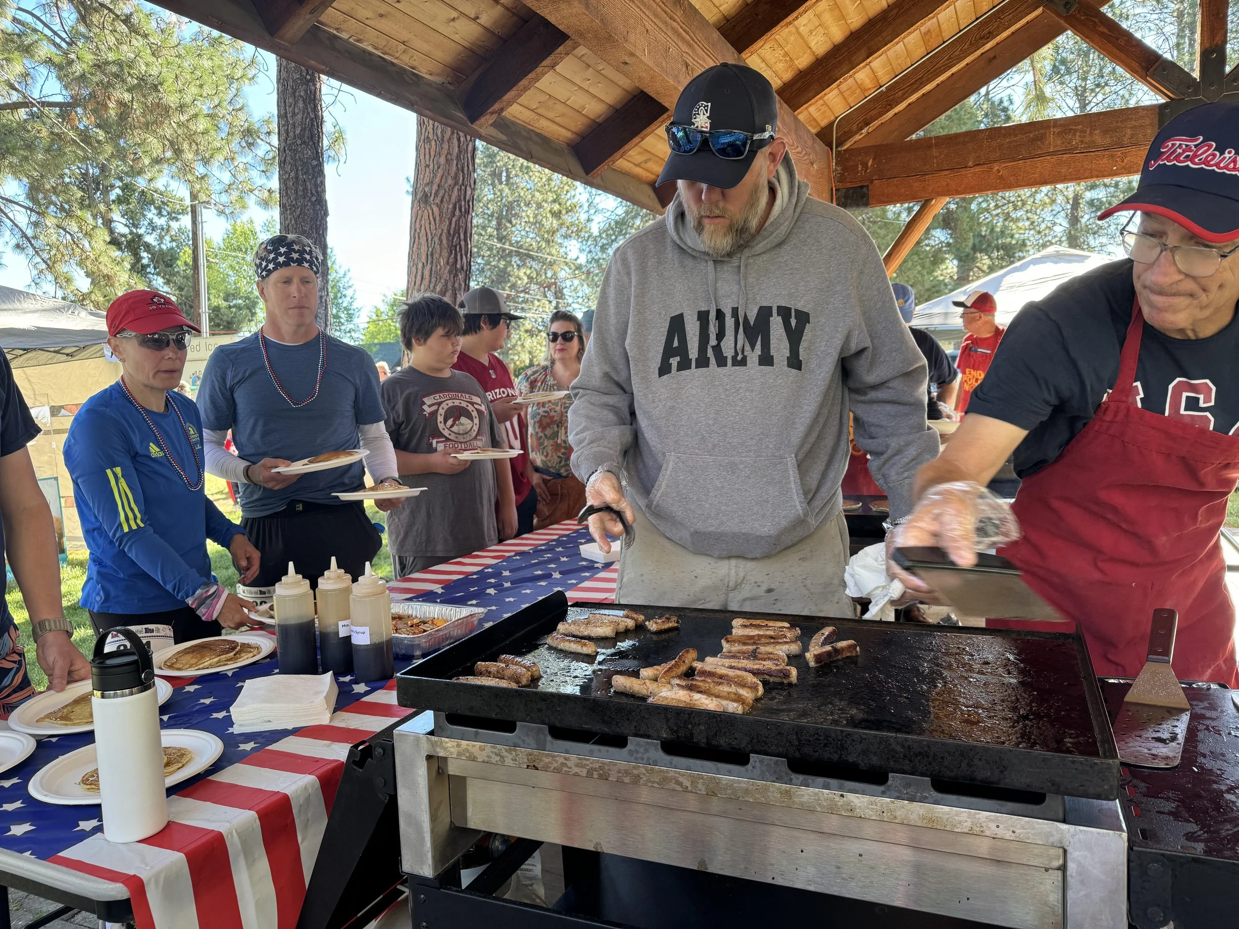People attending an outdoor pancake breakfast, grilling sausages on a large flat-top grill, with patriotic American flags and red, white, and blue decorations, under a wooden pavilion surrounded by trees.