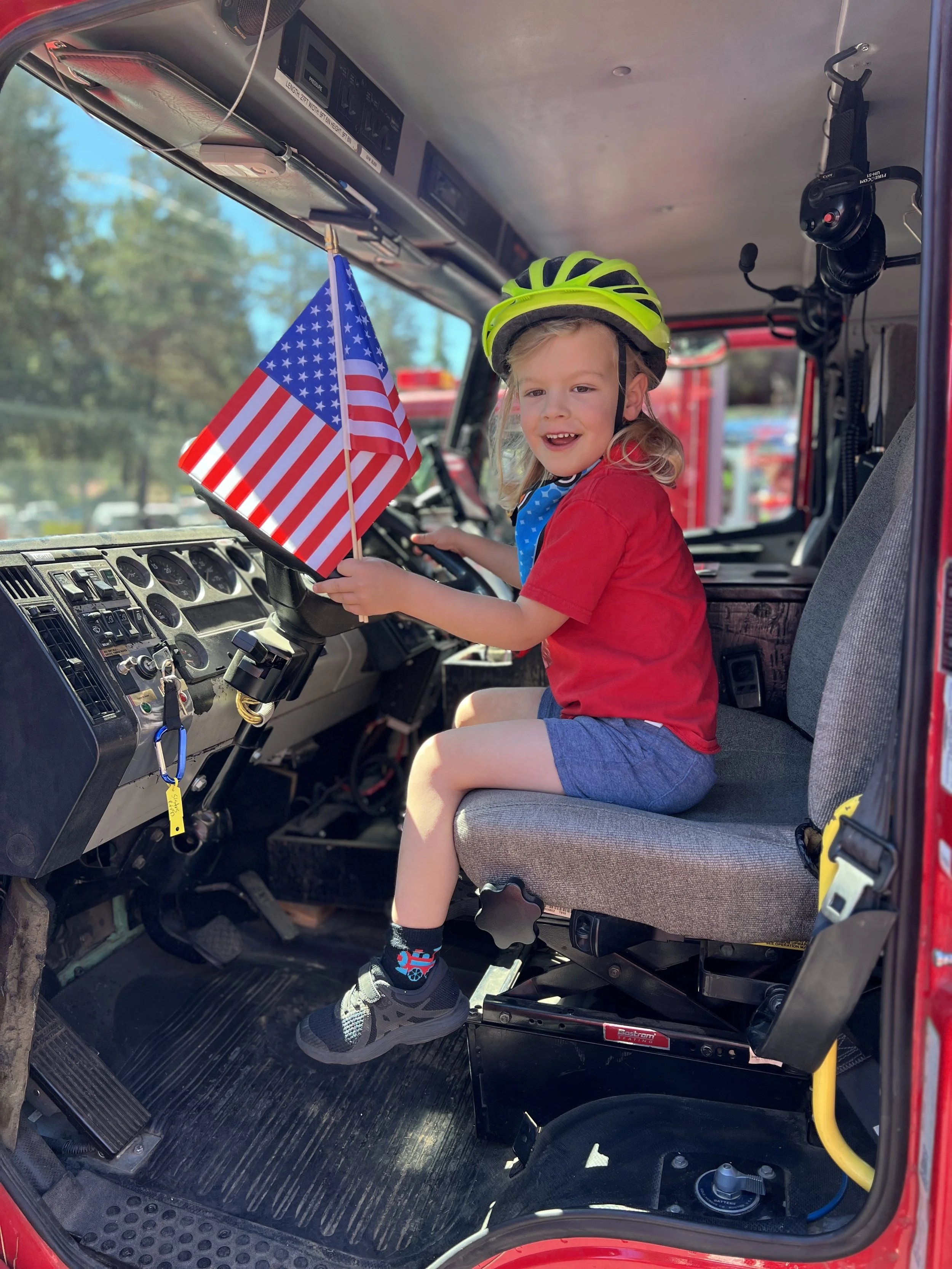 A young boy sitting in the driver's seat of a fire truck, wearing a neon green helmet, red shirt, and blue shorts, holding an American flag, with a smile on his face.