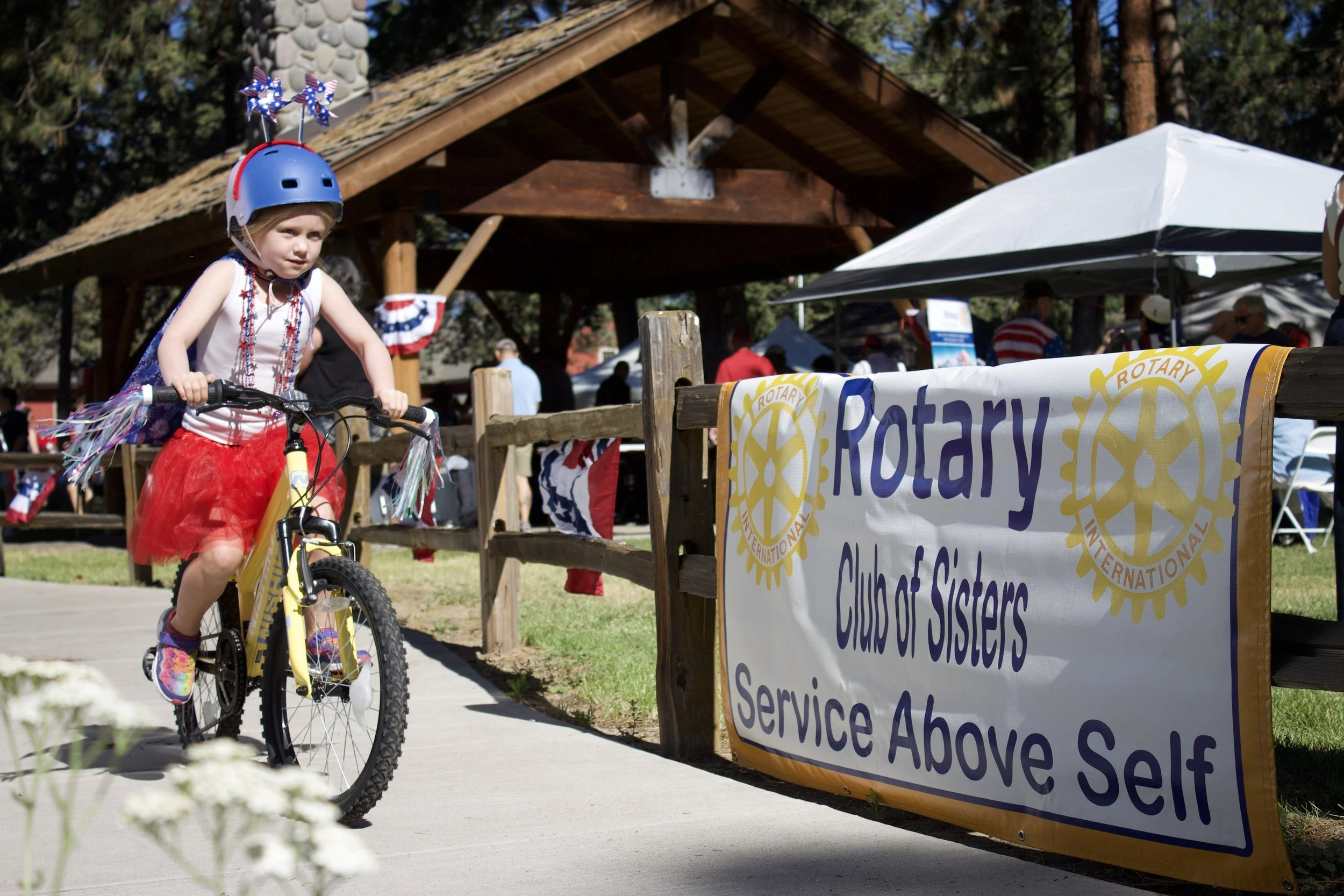 Young girl riding a yellow bicycle at an outdoor event with a Rotary Club banner in the foreground and a wooden pavilion with trees in the background.