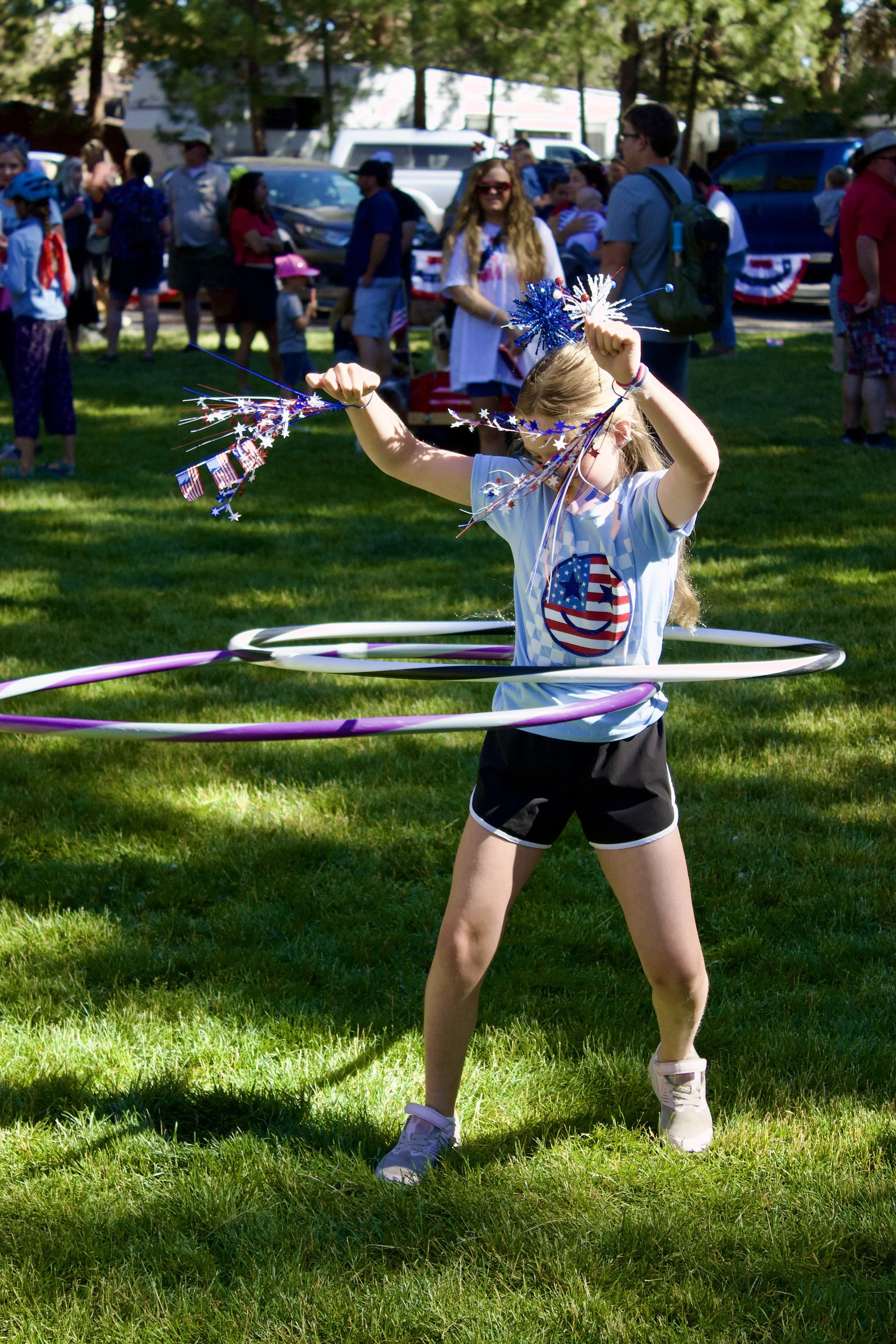 A young girl with blonde hair wearing a patriotic t-shirt, shorts, and sneakers standing on grass, hula hooping with red, white, and blue star- and flag-themed decorations during Sisters 4th Fest.