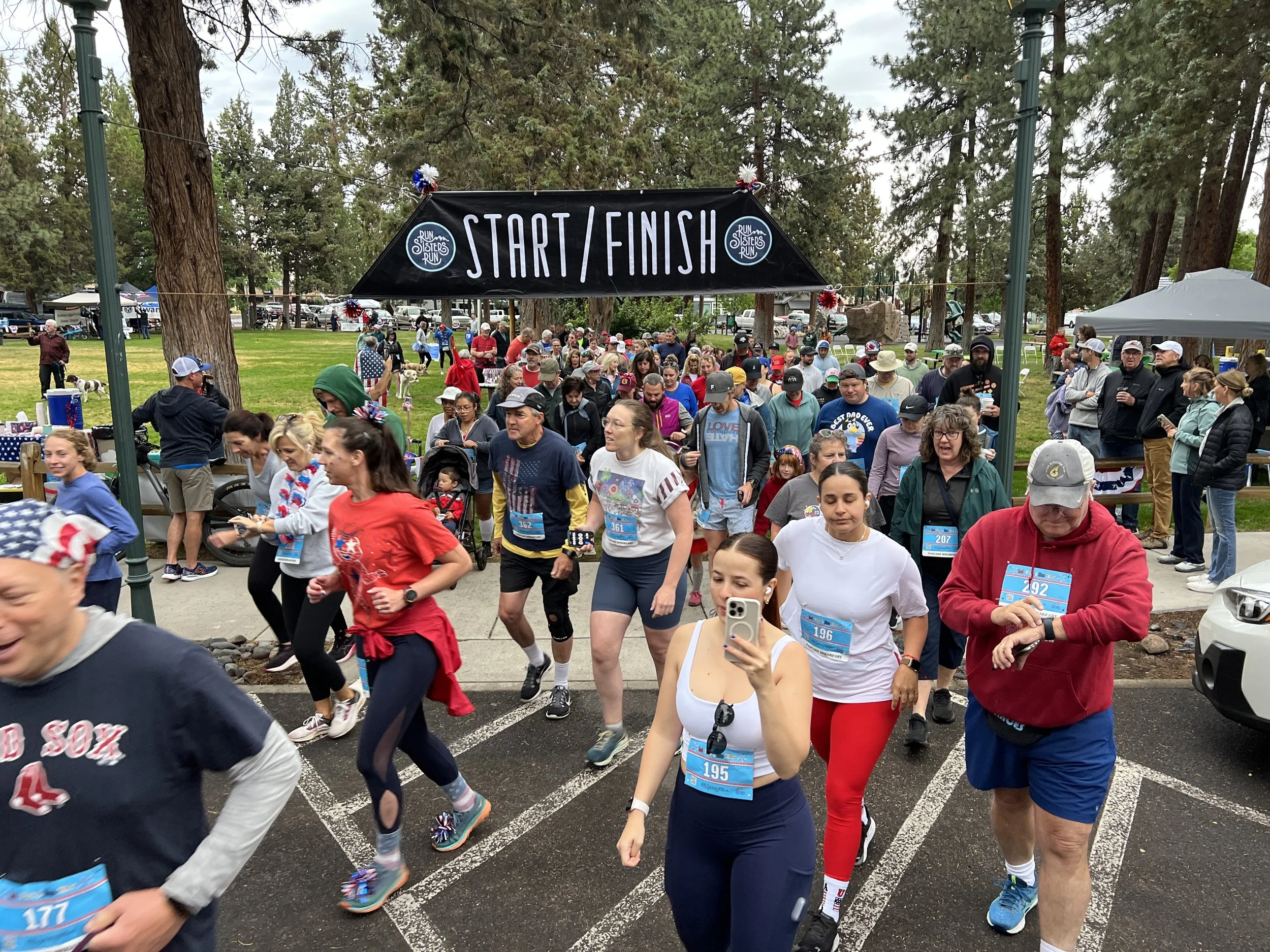 A crowd of people participating in Sisters 4th Fest's annual fun run, passing under a black banner that says 'START / FINISH' with the event name 'Run Sisters Run' shown on both sides of the text, in a park with large trees.
