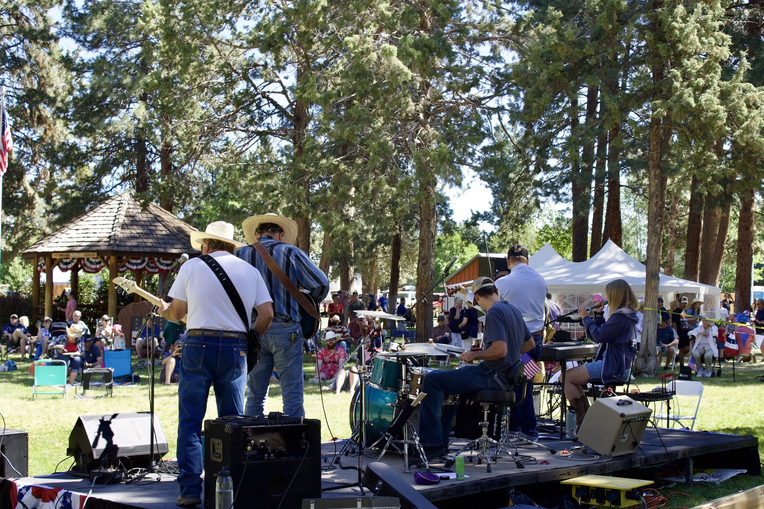 Musicians performing on an outdoor stage at a patriotic event, with many people watching in a park surrounded by tall trees and tents.