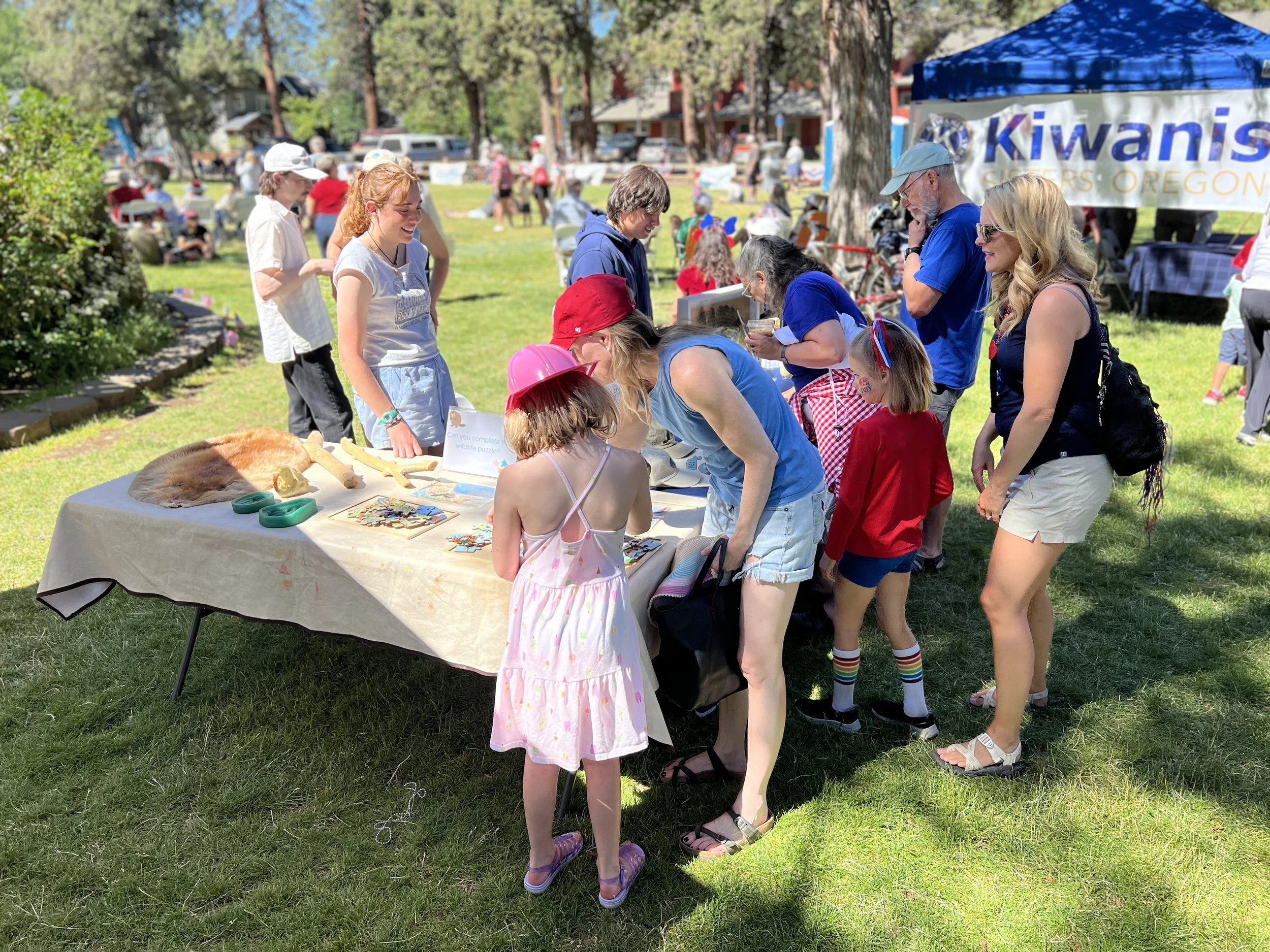 People gathered around a table at an outdoor event, with some children and adults examining items on display, under a sunny sky with trees and tents in the background.