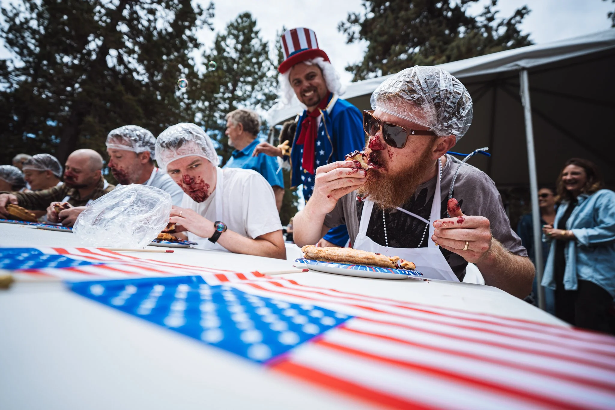 People participating in an American-themed pie eating contest outdoors, with some wearing hairnets, eating marionberry pie, and one person dressed as Uncle Sam with a tall striped hat and patriotic clothing.