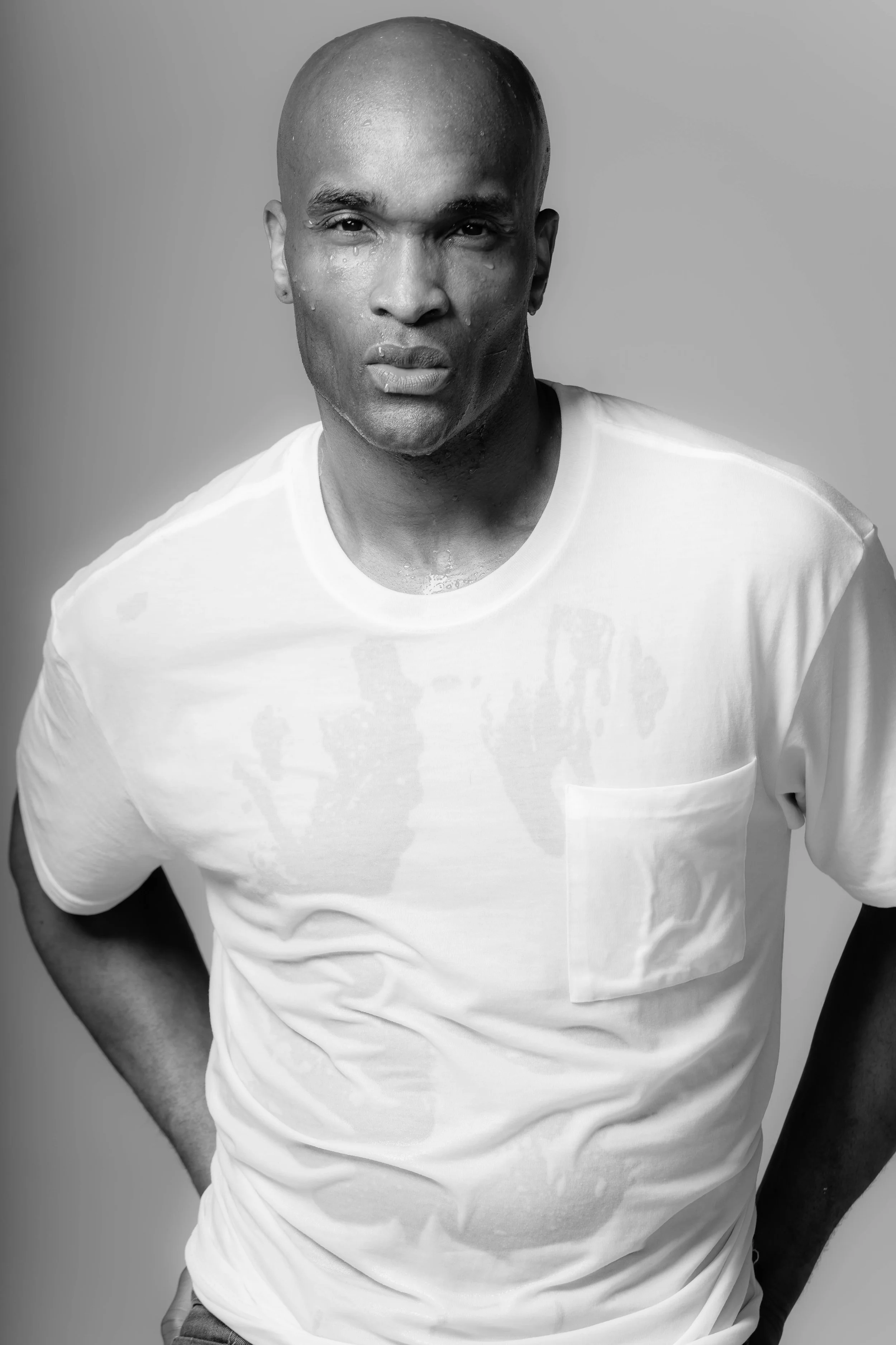 Black and white portrait of a man with a shaved head wearing a white t-shirt, standing against a plain background.
