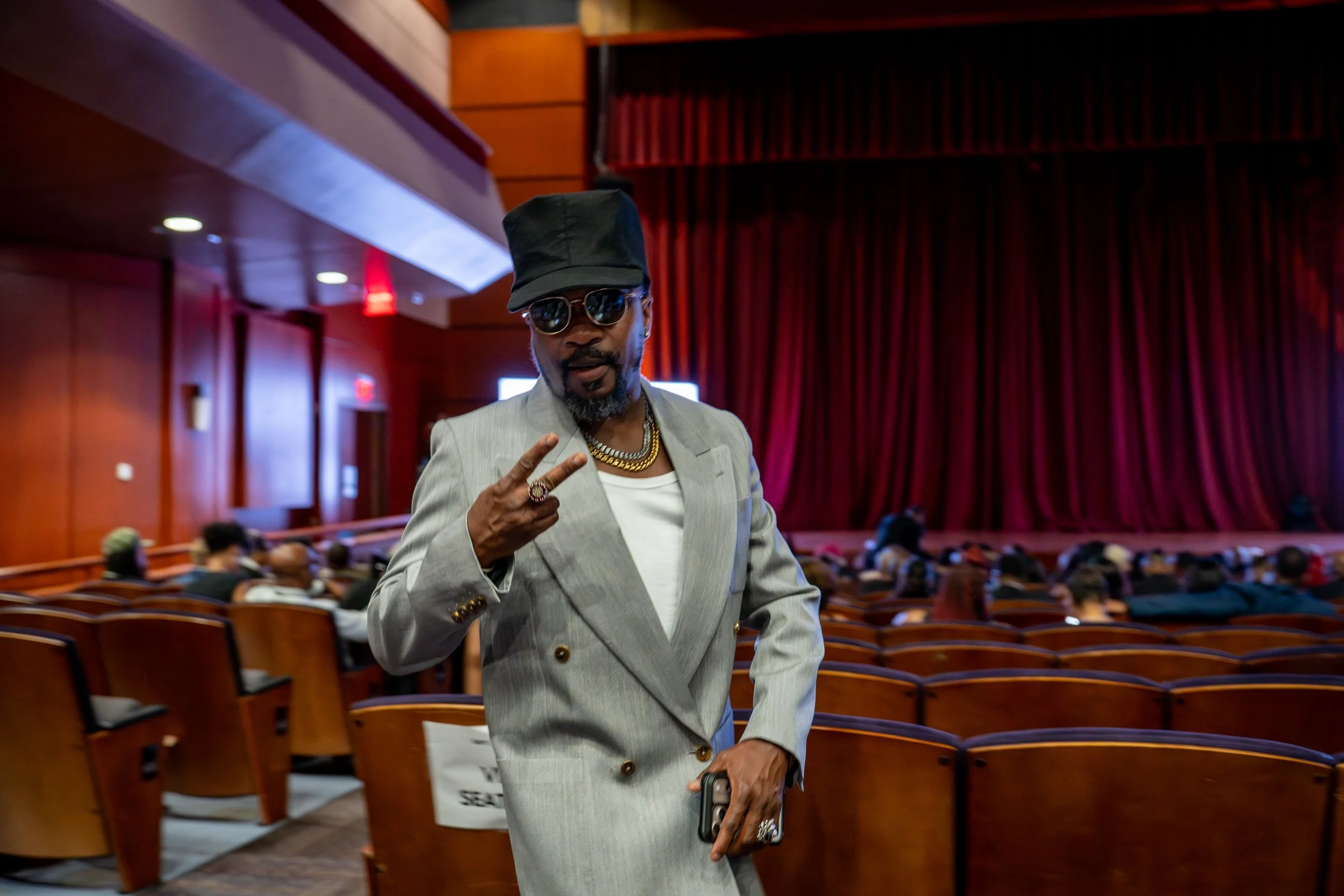A man dressed in a light gray suit with a white shirt, wearing dark sunglasses and a black hat, raising his hand in a peace sign inside an auditorium with red curtains and wooden paneling.