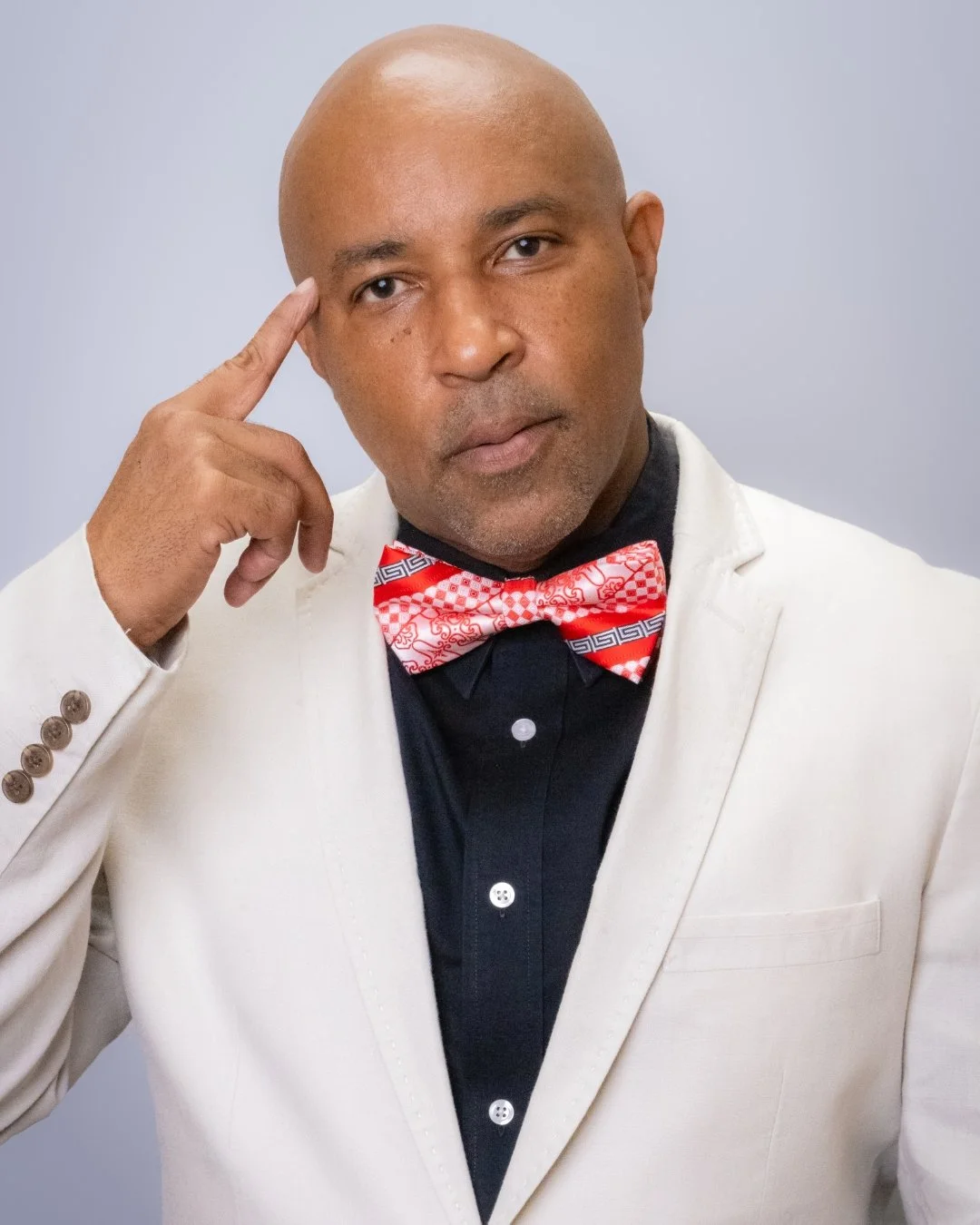 A man with a bald head, wearing a white suit jacket, black shirt, and a red and white patterned bow tie, is touching his temple with his right hand and looking at the camera.