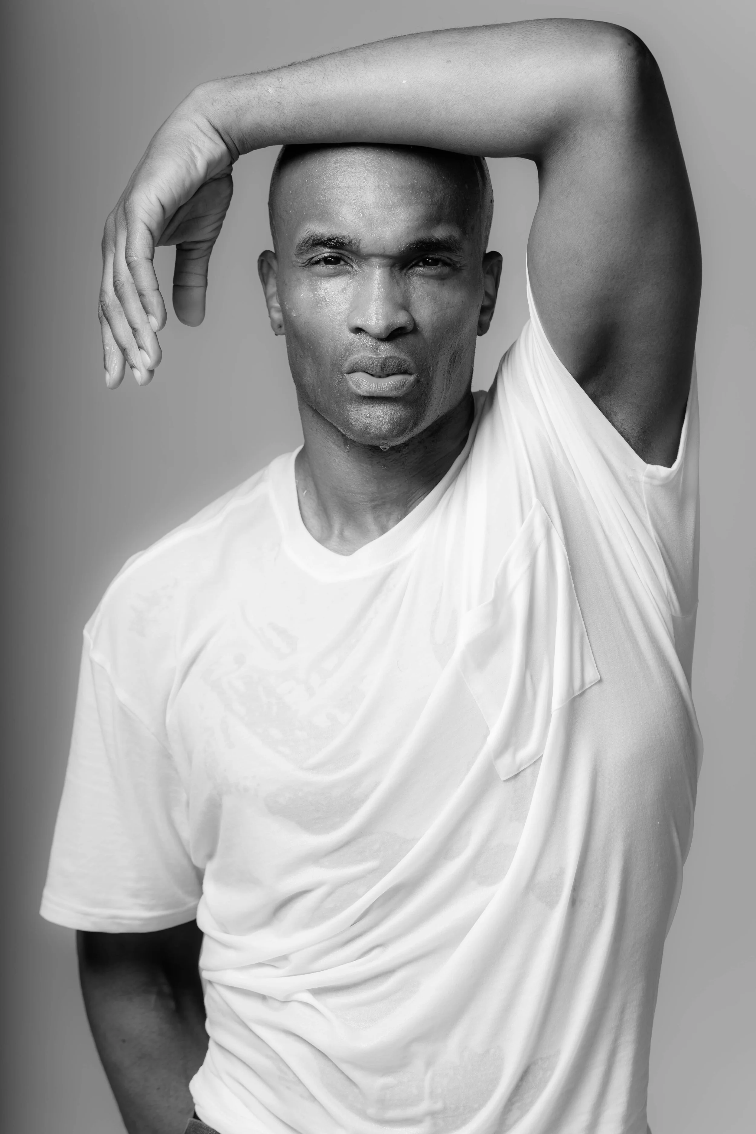 Black and white portrait of a man with a shaved head and muscular arm, wearing a white t-shirt, posing with his left arm raised and resting on his head, looking directly at the camera.