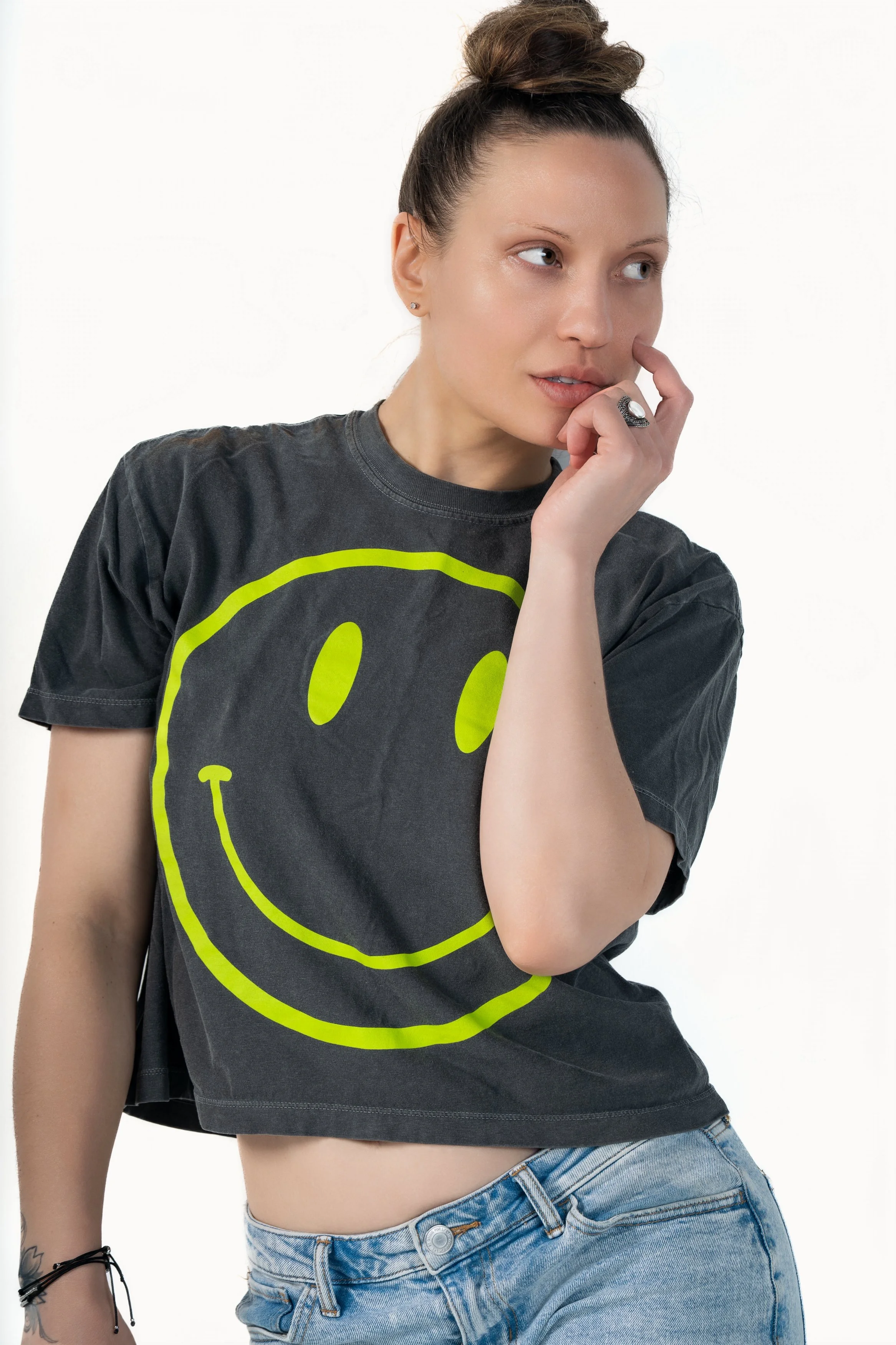 A young woman with light skin and brown hair styled in a bun wearing a black T-shirt with a neon yellow smiley face graphic and light blue jeans. She is touching her lips with her right hand and has a contemplative expression.