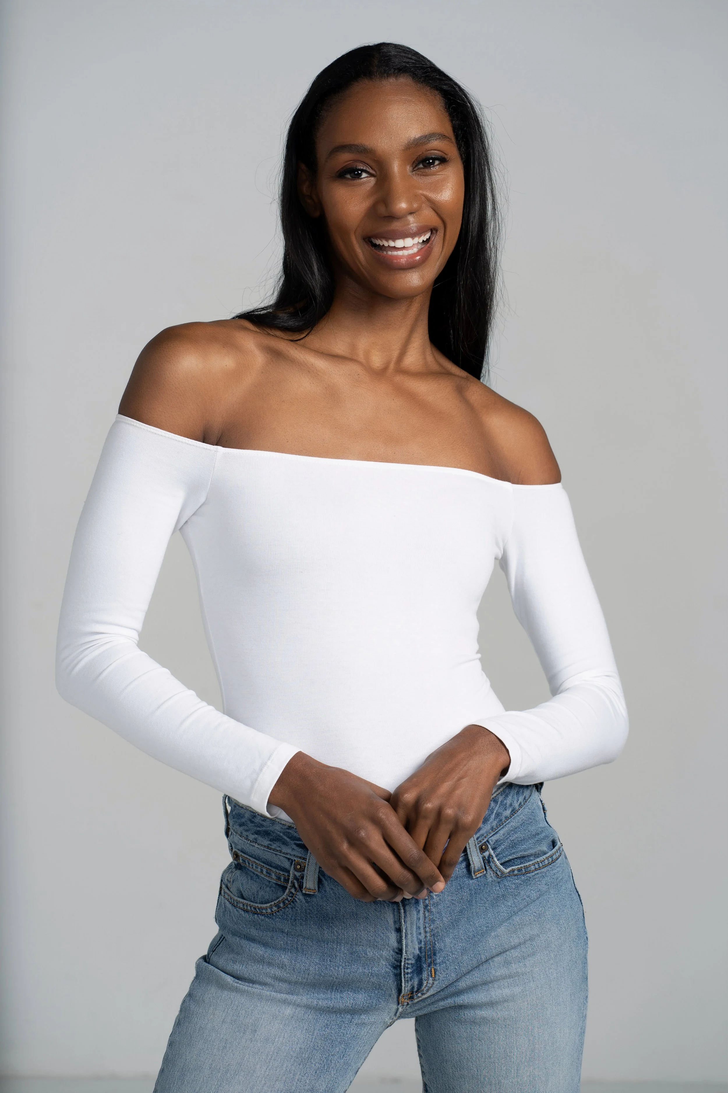A woman with medium-dark skin tone and long black hair, wearing a white off-the-shoulder top and blue jeans, standing against a plain light gray background.