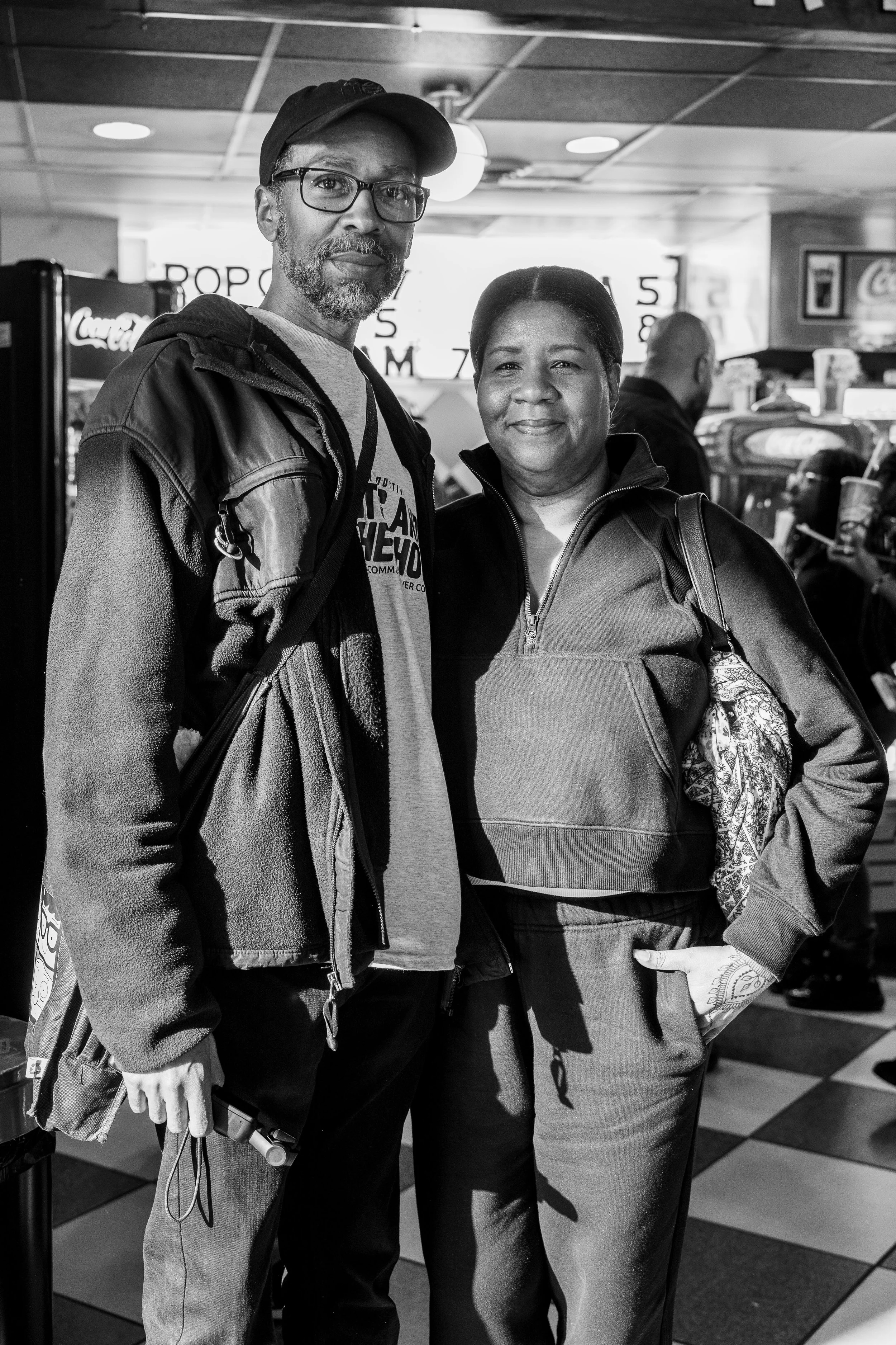 A man with glasses and a beard, and a woman with short hair, smiling at the camera, inside a store or mall, with signs for popcorn and beverages in the background.
