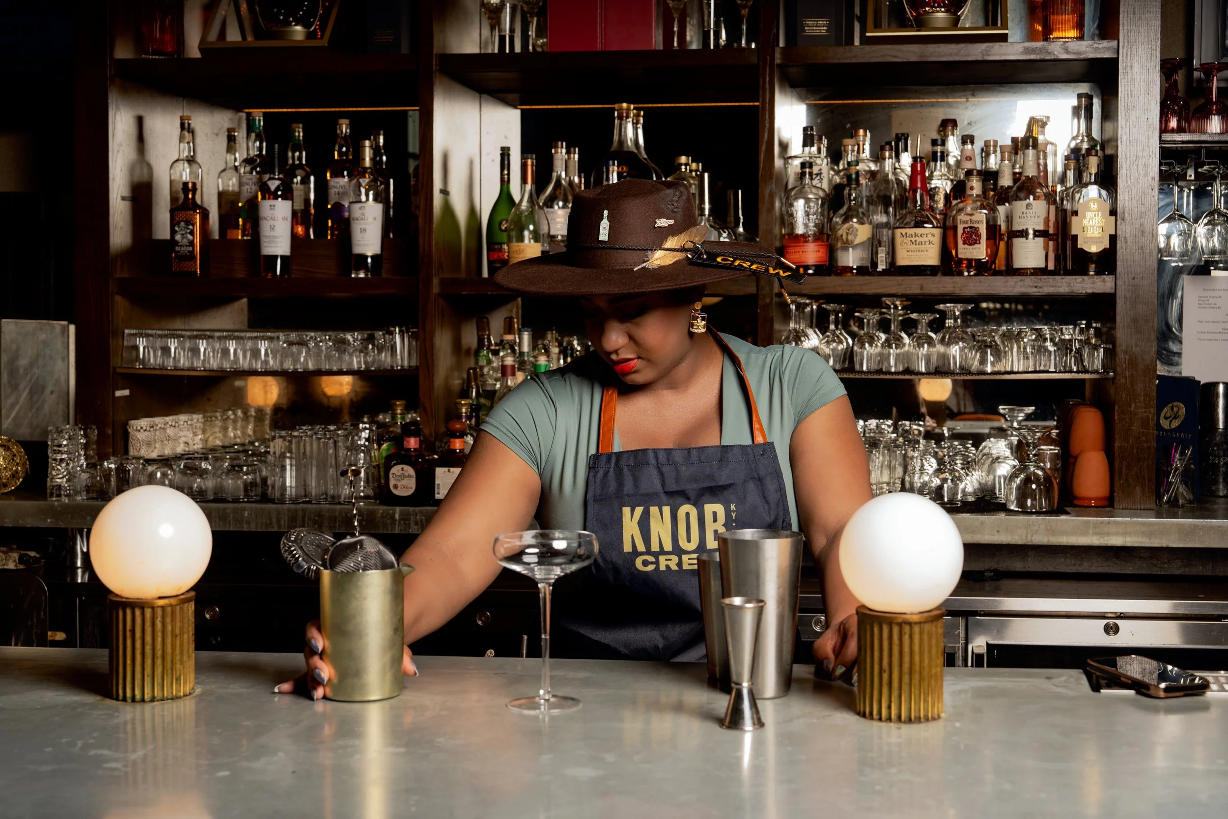 A female bartender with a wide-brimmed hat behind a bar, preparing cocktails. The bar is decorated with bottles of liquor on shelves in the background and illuminated spherical lights on the counter.
