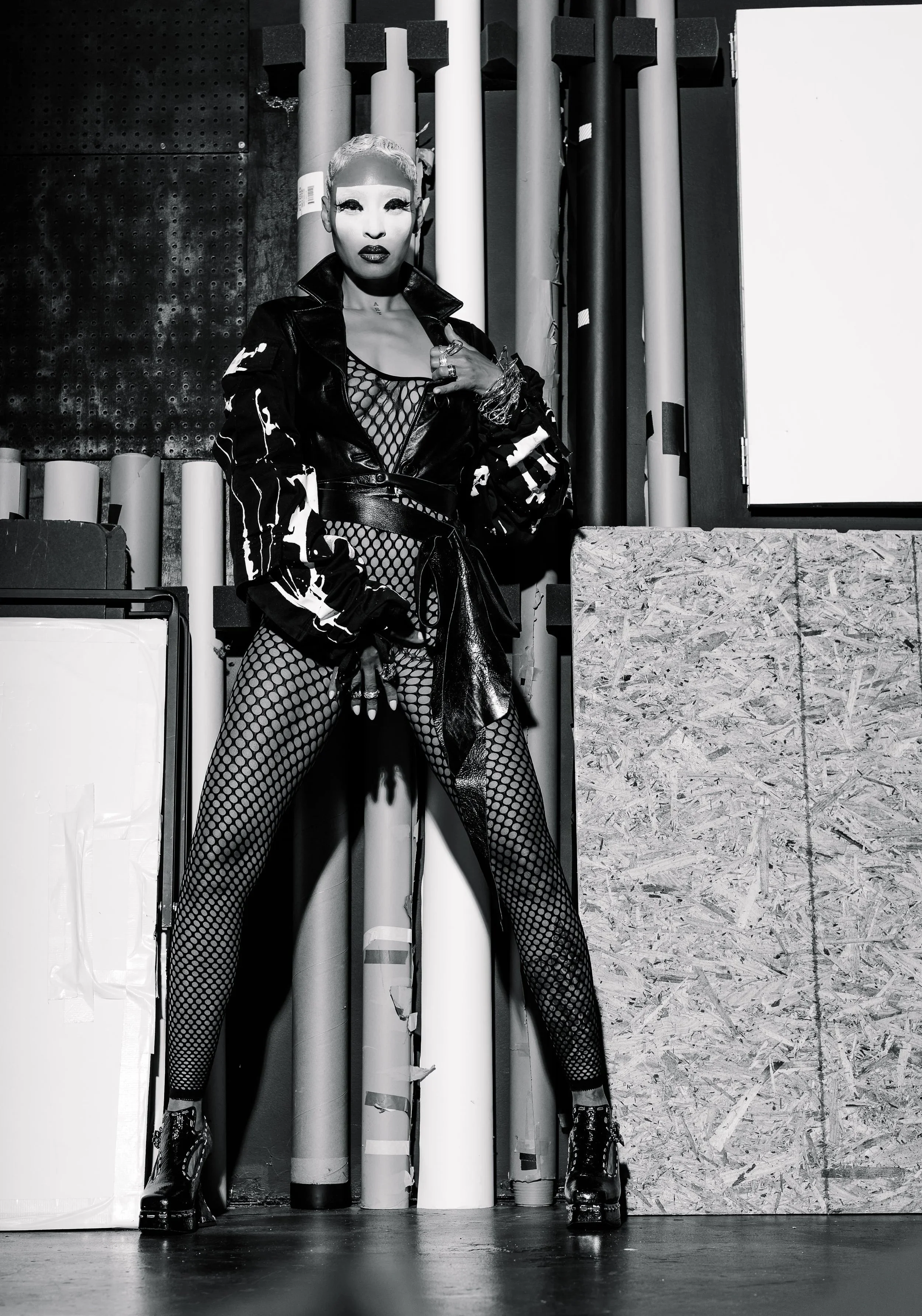 A woman in black fishnet tights, a leather jacket, and platform boots posing with one hand on her chest and in front of a background with cylindrical pipes and textured surfaces in grayscale.