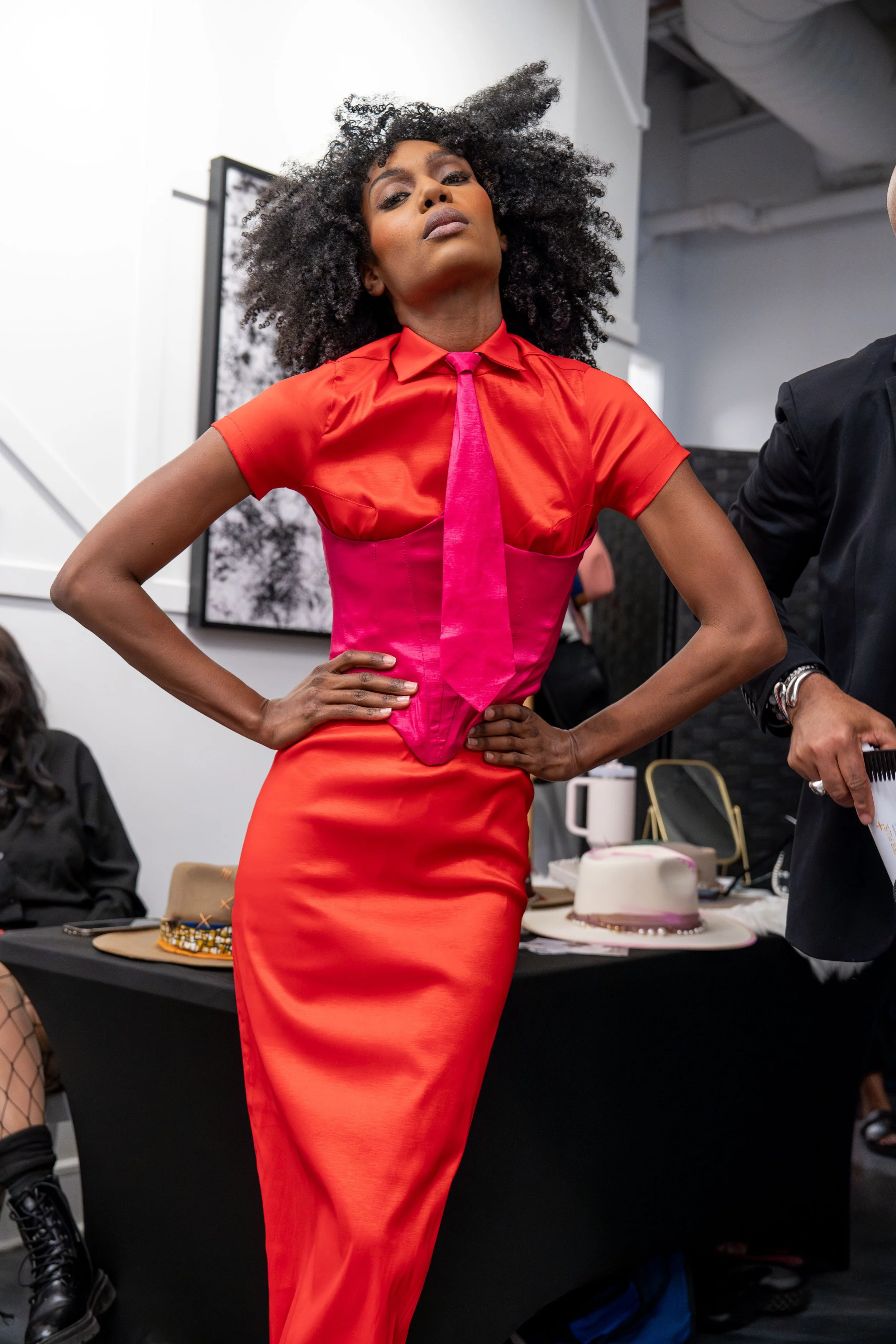 A woman in a vibrant red dress with pink accents poses confidently with her hands on her hips in an indoor setting. Behind her, a table displays various hats and jewelry, and a woman seated in the background.