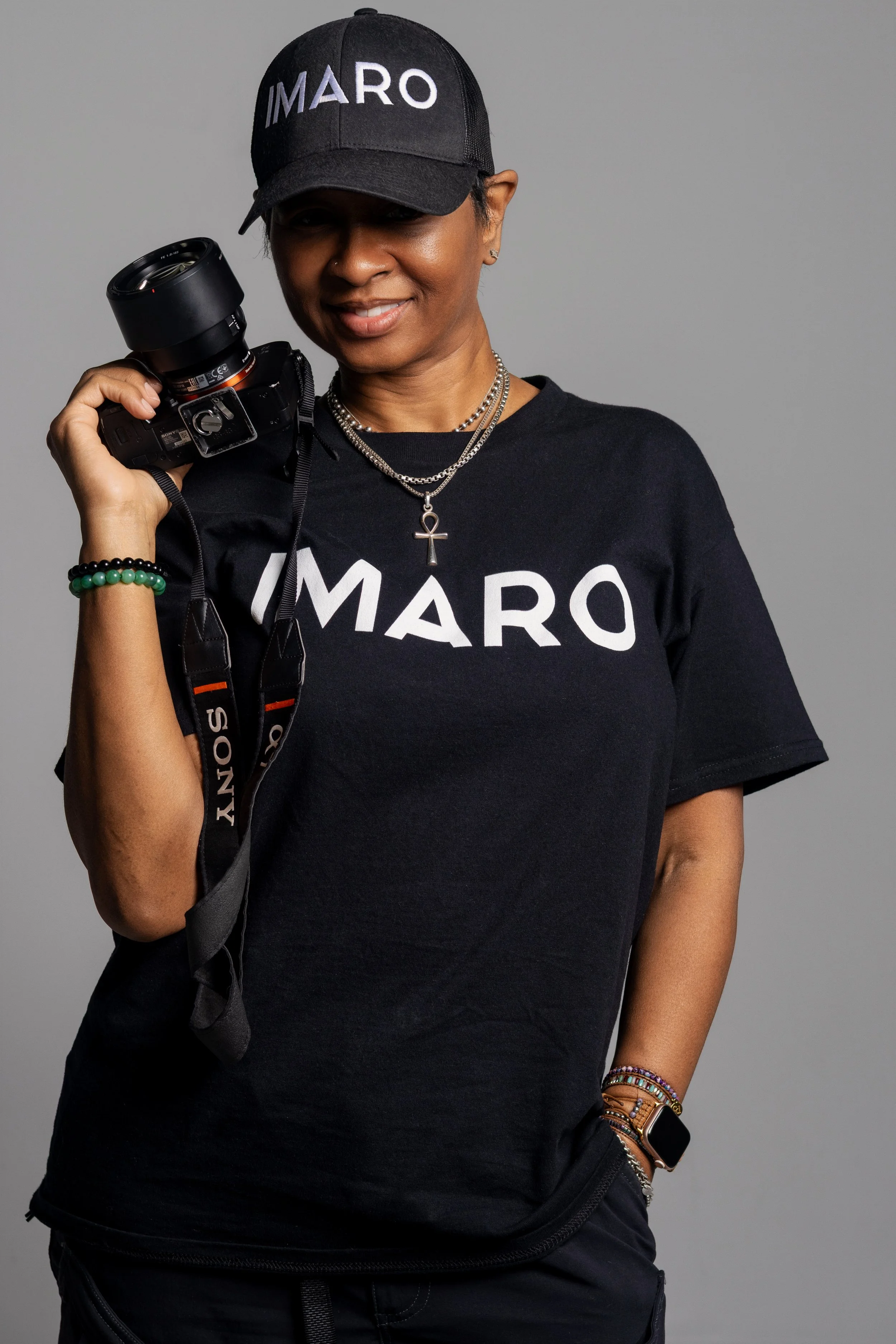 A woman smiling, holding a camera camera with a strap, wearing a black cap and matching black T-shirt with the word 'IMARO' printed on it, and accessorized with necklaces, bracelets, and a watch, standing against a plain gray background.
