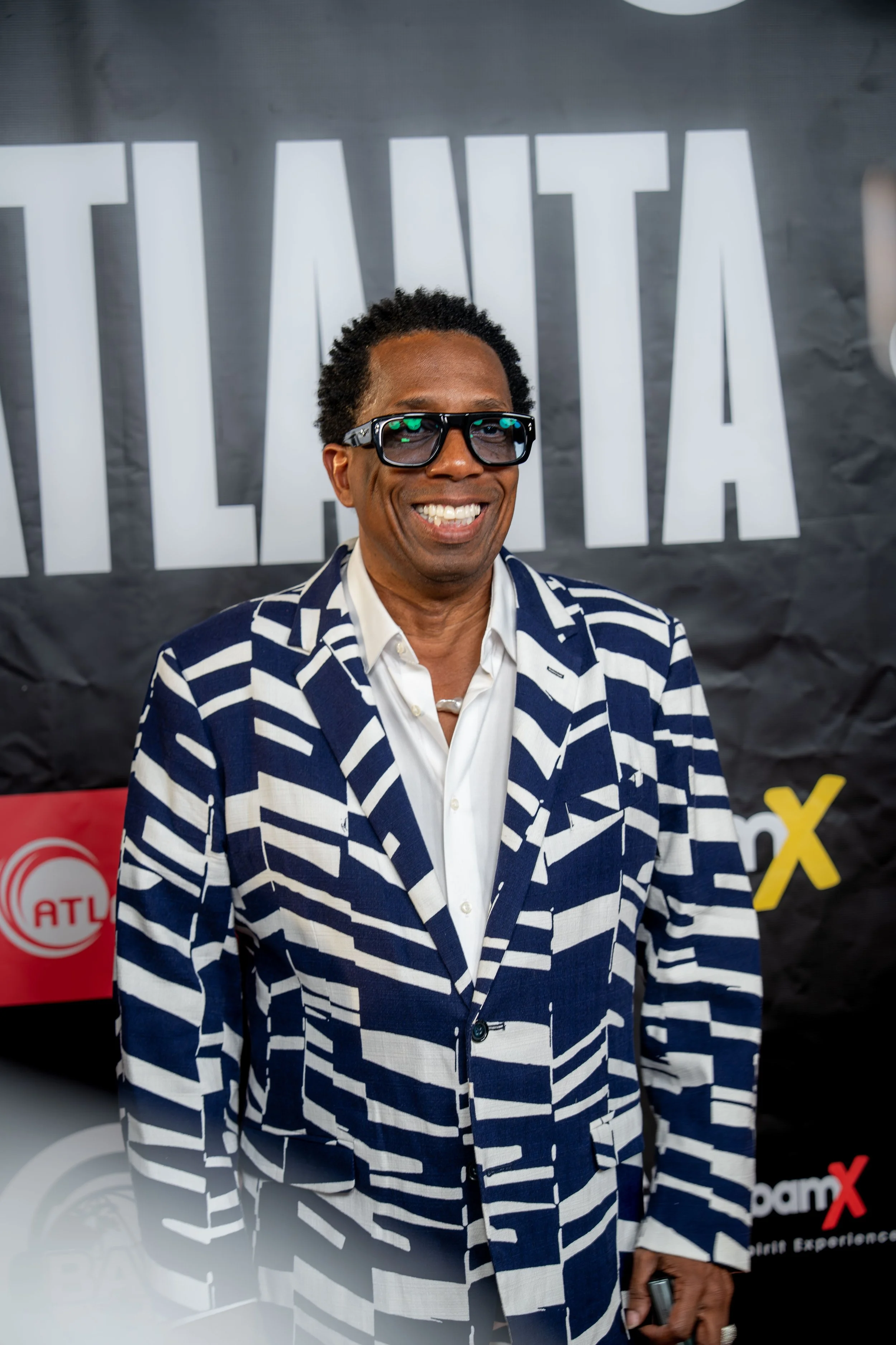 A smiling man wearing a navy and white striped blazer, white shirt, and sunglasses standing in front of a black background with white and yellow logos, including 'ATLI' in a red circle and 'X' in red and black.