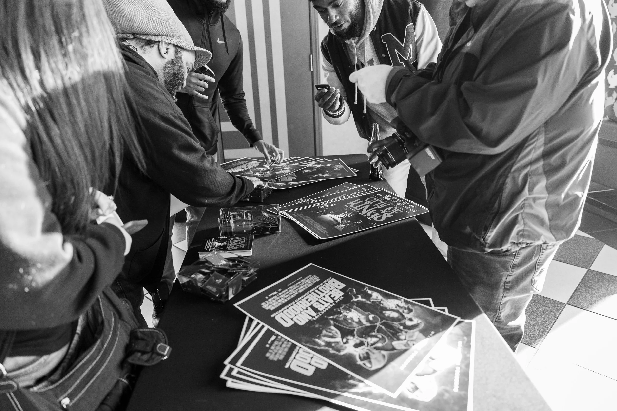 Group of people gathered around a table with posters promoting an event or concert, looking at and discussing the posters.