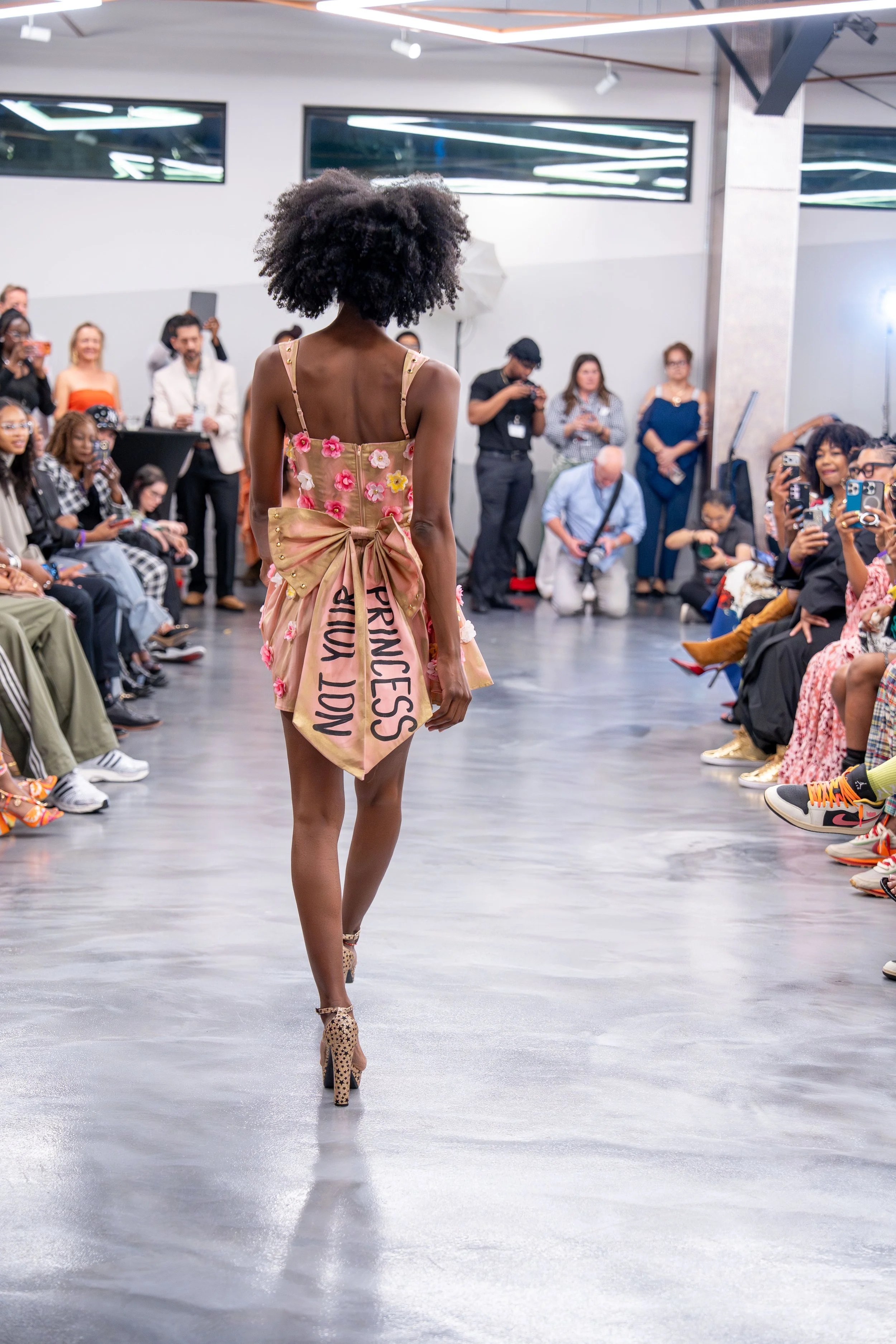 A female model walking down the runway at a fashion show, wearing a beige dress with pink and white flowers and a bow at the back, surrounded by an audience taking photos.