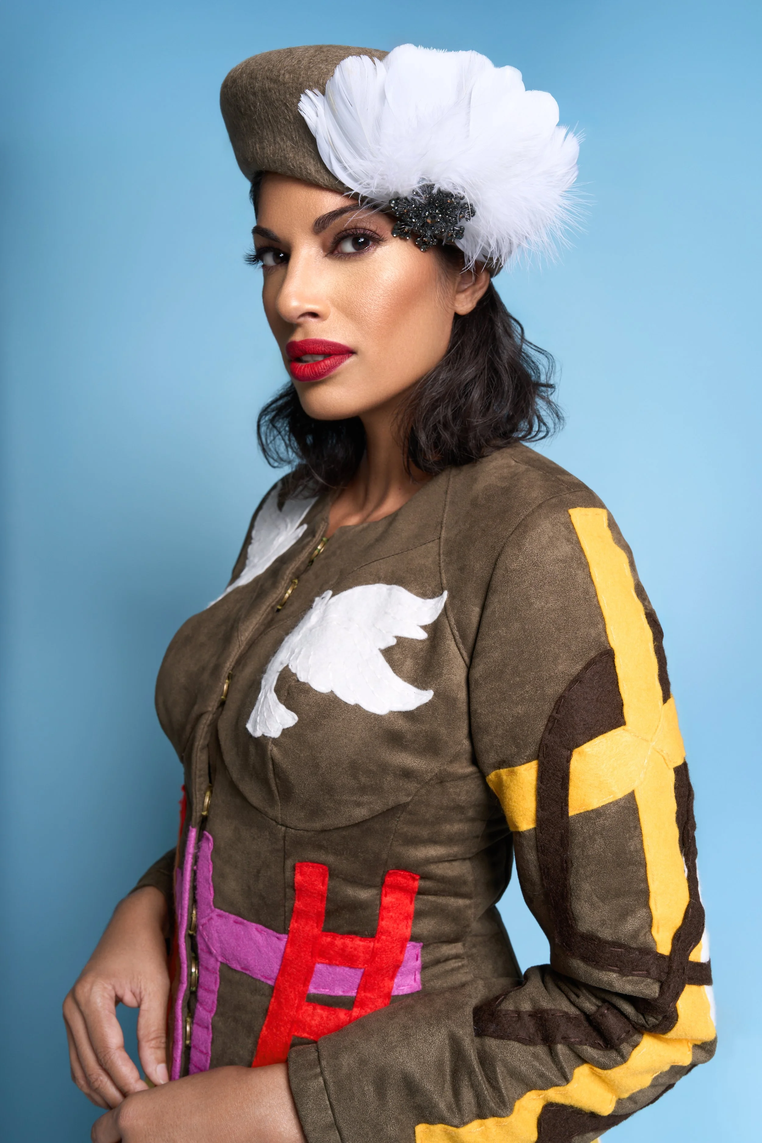 A woman wearing a beige military-style hat decorated with a large white feather and black embellishments, and a tan jacket with colorful patches and embroidery, against a plain blue background.