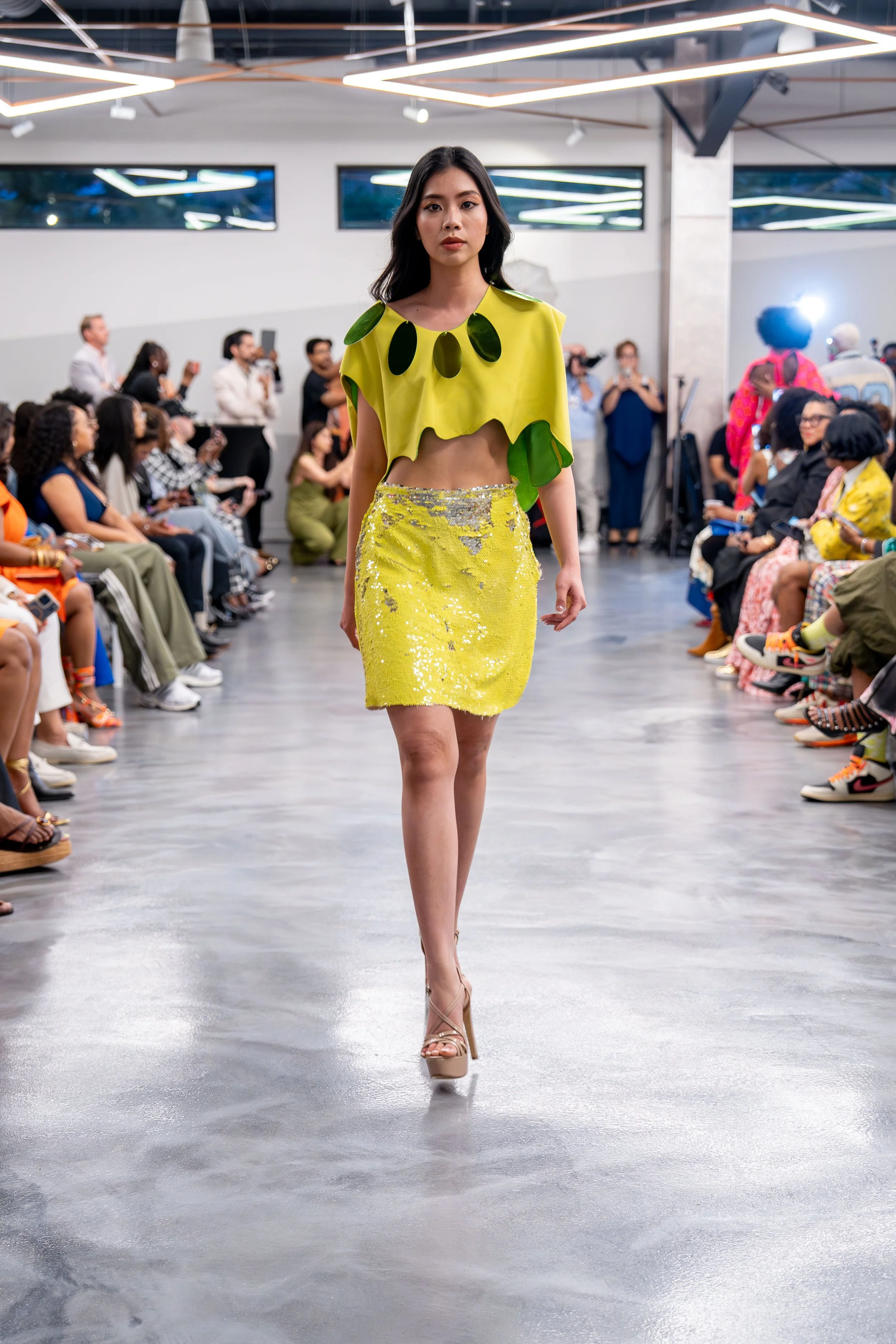 A fashion runway show featuring a model walking in a designer outfit. The model wears a bright yellow two-piece outfit with a cropped top and a shimmering skirt, paired with high heels. The audience is seated on either side of the runway, attentively watching the show. The venue has a modern interior with geometric lighting fixtures.